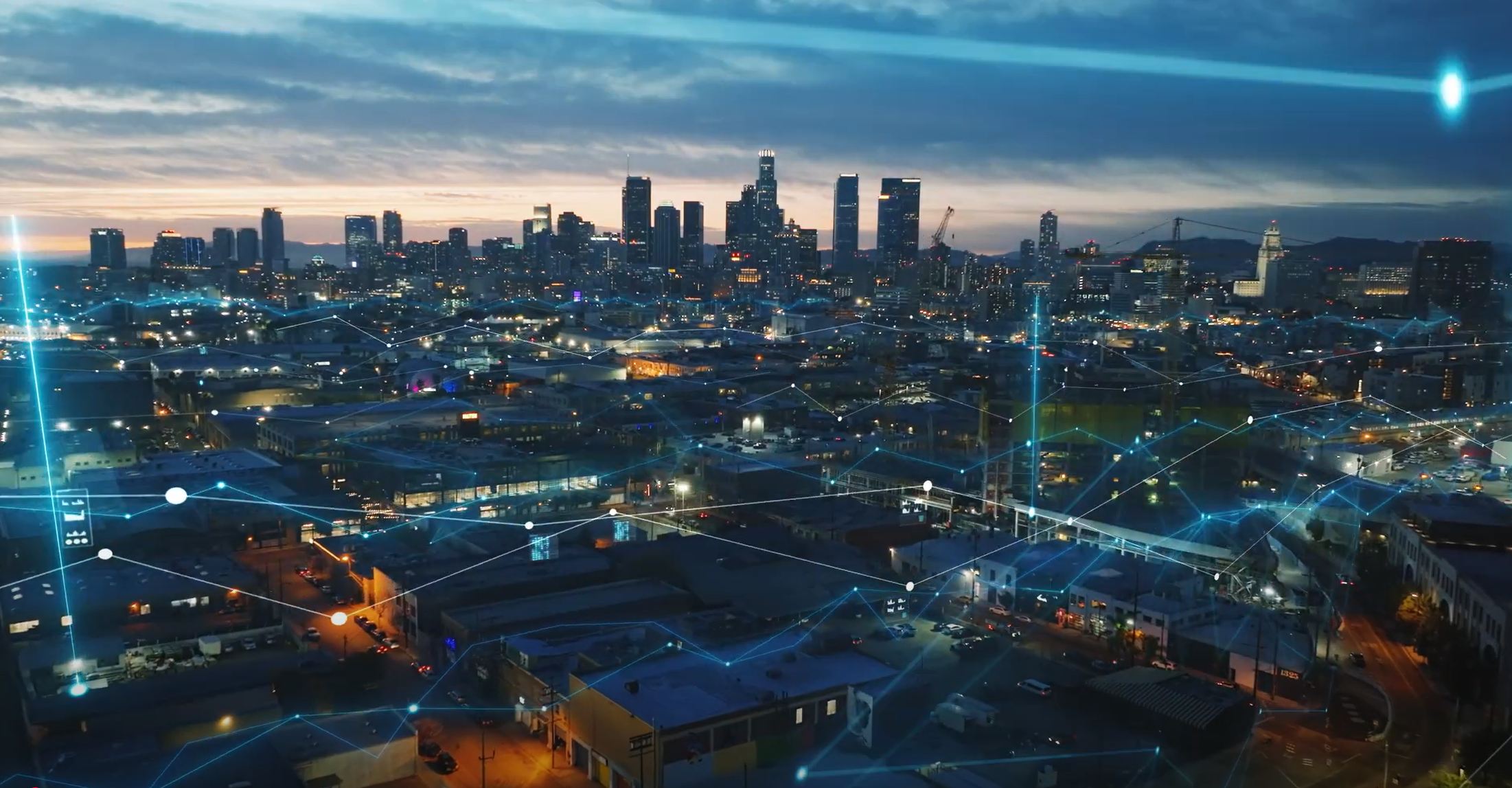 A cityscape at dusk with illuminated buildings and streets, overlaid with digital network lines, suggesting connectivity. - CSA Catapult