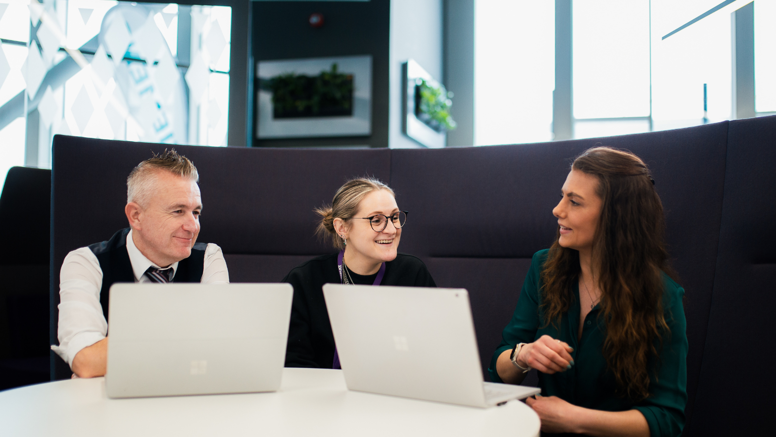 Three colleagues sit around a white table, each with a laptop in front of them. They are engaging in a conversation, smiling, and appear to be in a modern office setting with plants and geometric wall designs in the background. - CSA Catapult