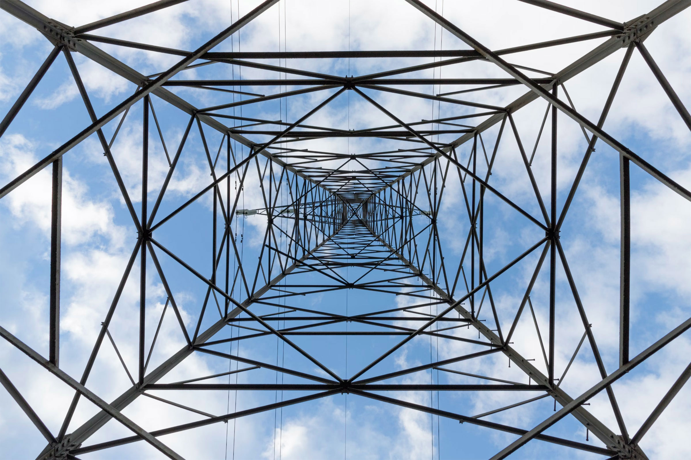 A symmetrical view looking up inside a metal electricity transmission tower against a blue sky with scattered white clouds. The crisscrossing metal beams form a geometric, tunnel-like pattern. - CSA Catapult