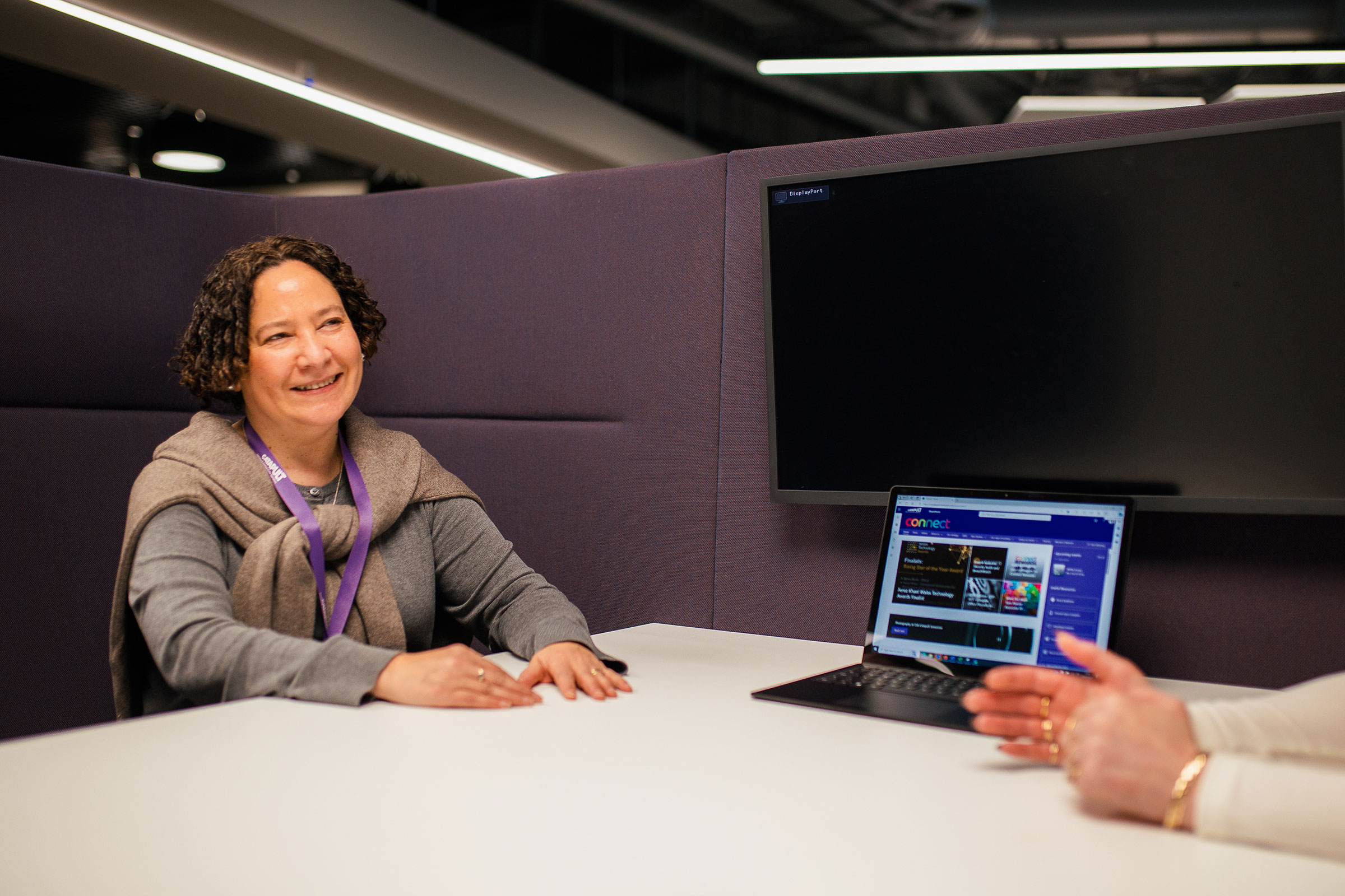 A woman sits at a table, smiling, and facing another person whose hands are visible. A laptop on the table displays a website. The setting appears to be an office with a modern design and purple partitions. - CSA Catapult