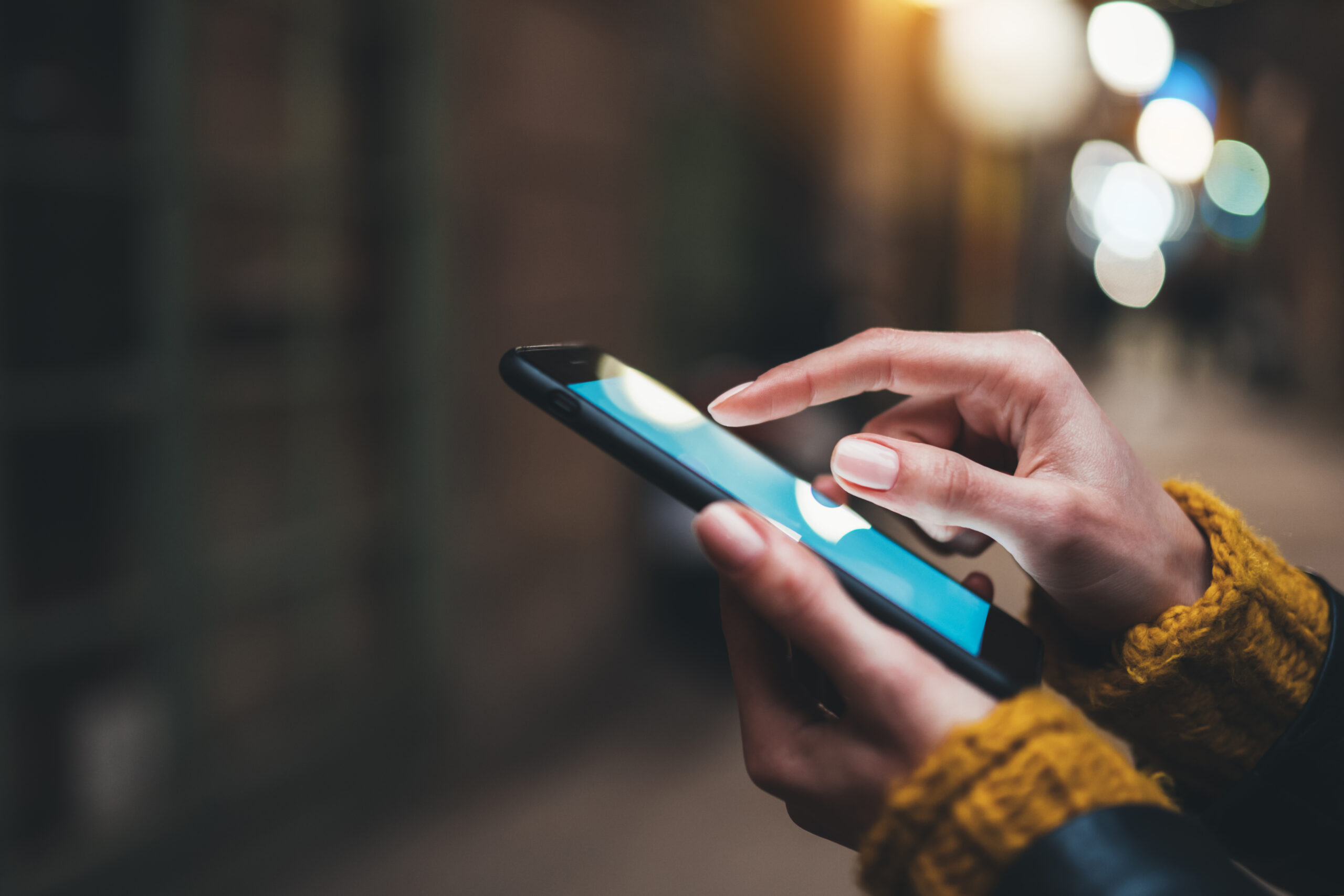 Close-up of a person’s hands using a smartphone. The individual has light-colored nail polish and is wearing a yellow sweater with a dark jacket. The background is blurred, featuring city lights and buildings, giving the scene a warm, evening ambiance. - CSA Catapult