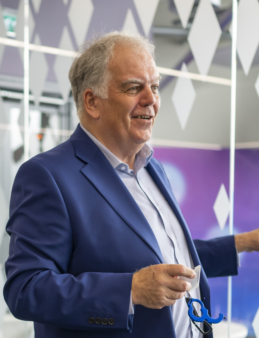 A man wearing a blue blazer and light blue shirt stands in an office environment. He is holding a pair of glasses in his left hand, looking to the side as if engaging with someone off-camera. The background features elegant geometric patterns. - CSA Catapult