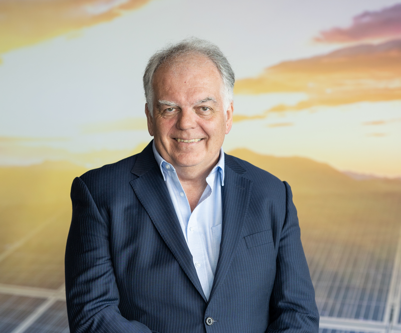 A smiling man with gray hair stands in front of a backdrop featuring solar panels and a sunset sky. He is dressed in a dark suit and has his hands loosely clasped in front of him, embodying a sense of serene confidence. - CSA Catapult