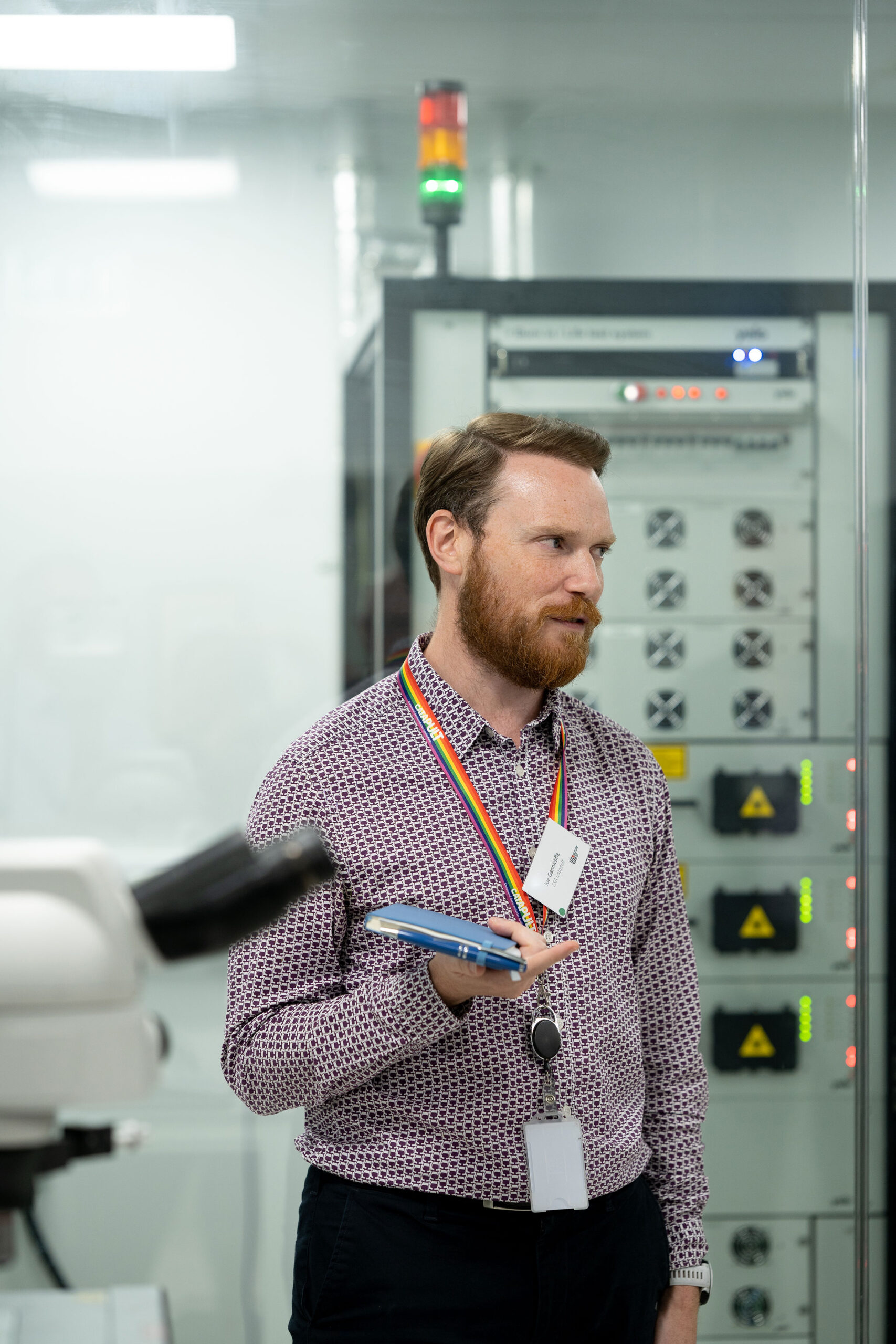 A bearded man stands in a high-tech laboratory, clutching a blue notebook and wearing a colorful lanyard with an ID card. Dressed in a patterned shirt, he is surrounded by scientific equipment and control panels. - CSA Catapult