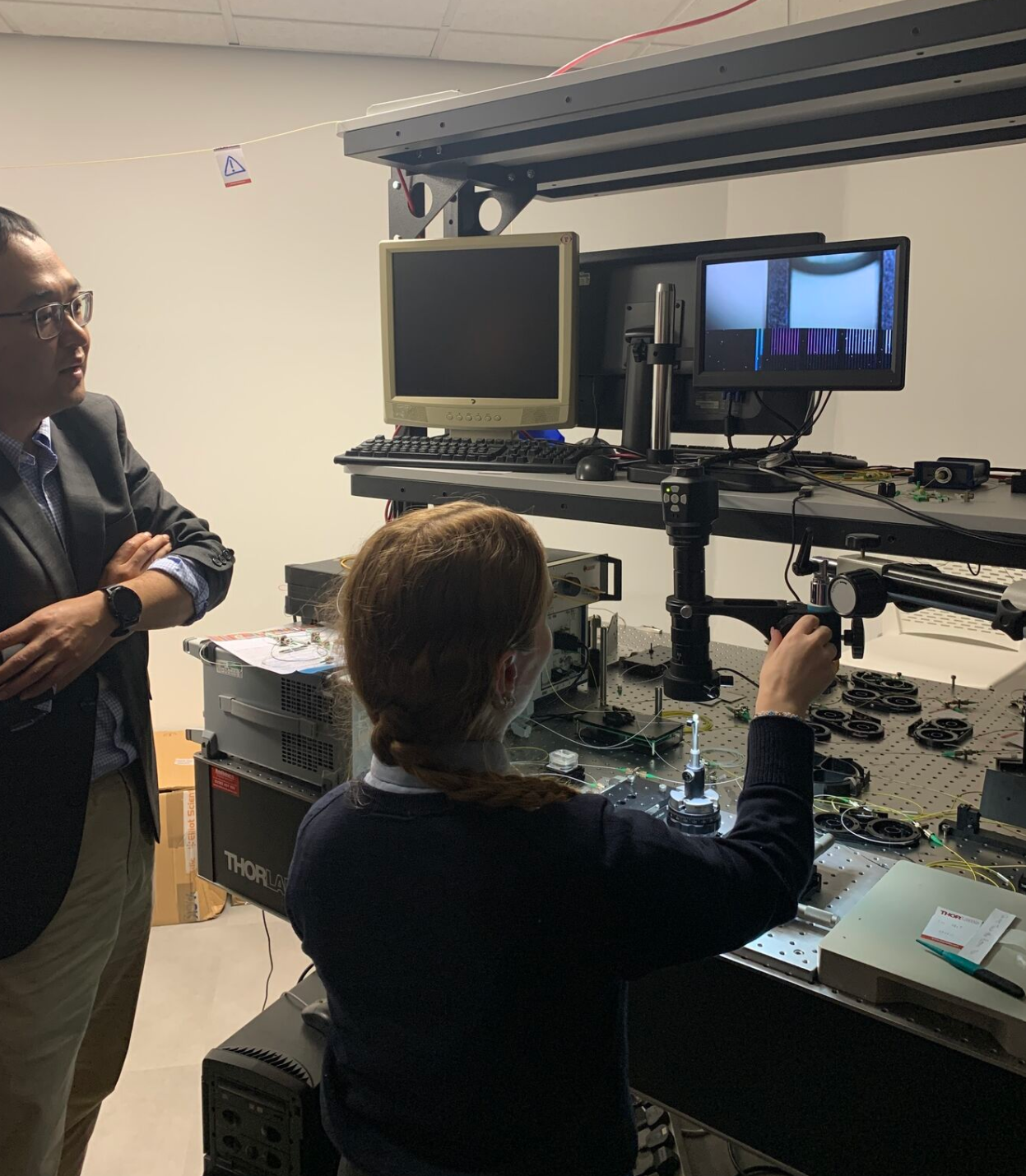 A man in a dark blazer and glasses observes as a woman in a ponytail operates a complex workstation with multiple monitors and electronic equipment. They are in a lab setting with specialized technical devices around them. - CSA Catapult