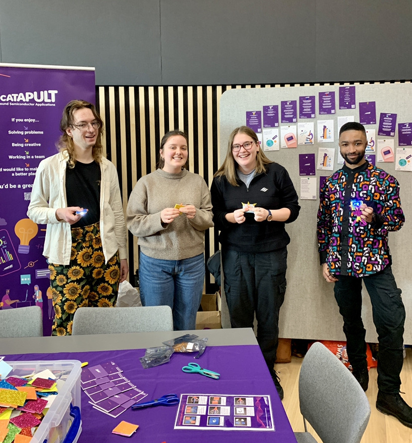 Four people stand side by side, smiling and holding small crafts, in front of a table with various crafting materials and tools. Behind them is a tall banner with text and a wall display of colorful paper items forming the letter 