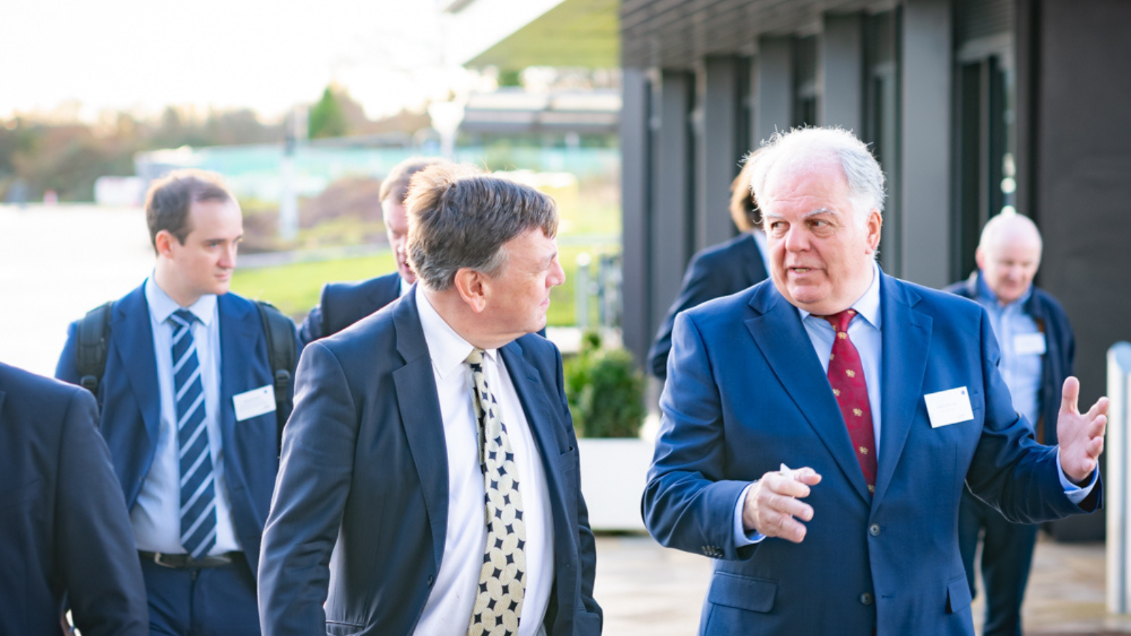 A group of men in business attire are walking and conversing outside. The man on the right, wearing a blue suit and a tie, is gesturing with his hand while speaking to another man in a similar suit. Other people in suits are visible in the background. - CSA Catapult