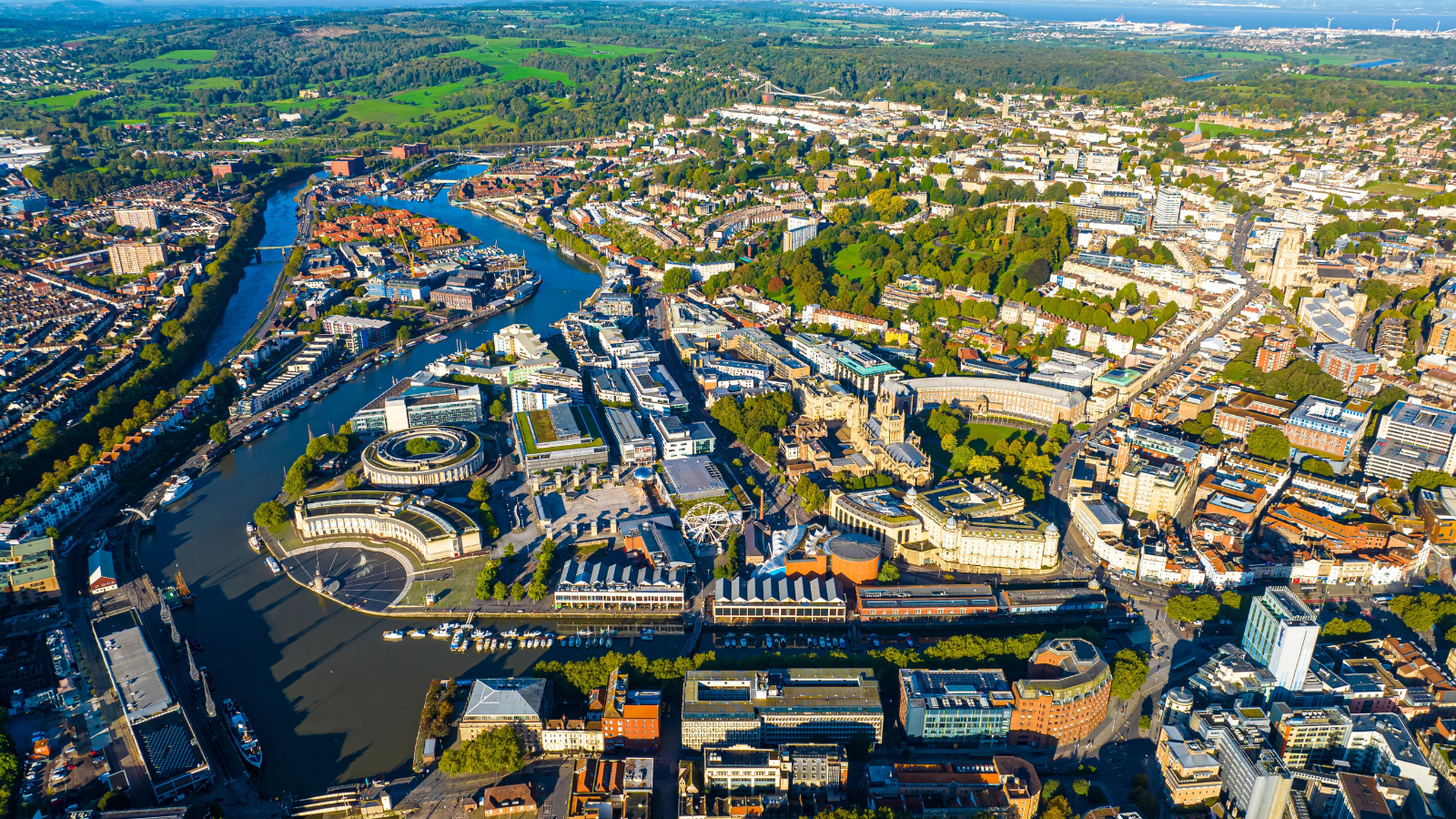 Aerial view of a sprawling city with a river cutting through its center. The city features a mix of modern and historic buildings, lush green spaces, and a winding network of streets and bridges. Surrounding areas consist of suburbs and patches of greenery. - CSA Catapult