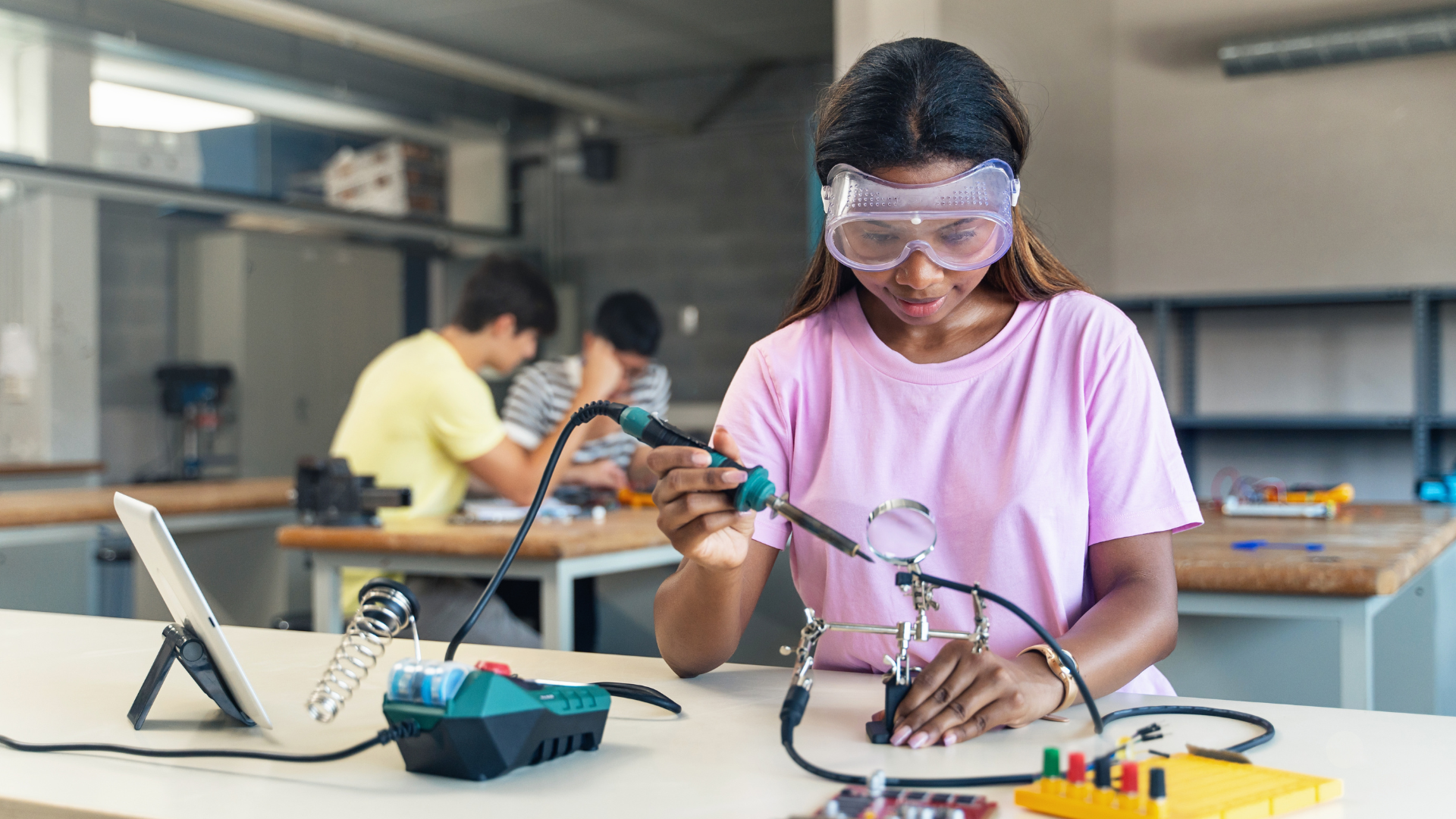 A young woman wearing safety goggles solders electronic components at a workstation. She is focused on her task, with tools and equipment spread out on the table. In the background, two individuals work on similar projects in a workshop setting. - CSA Catapult