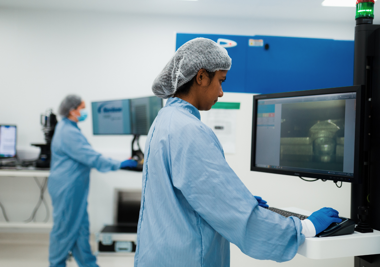 Two lab technicians in blue protective suits work at computer stations in a bright, clean laboratory. One technician operates a computer connected to a large machine, while the other works on a different piece of equipment in the background. - CSA Catapult