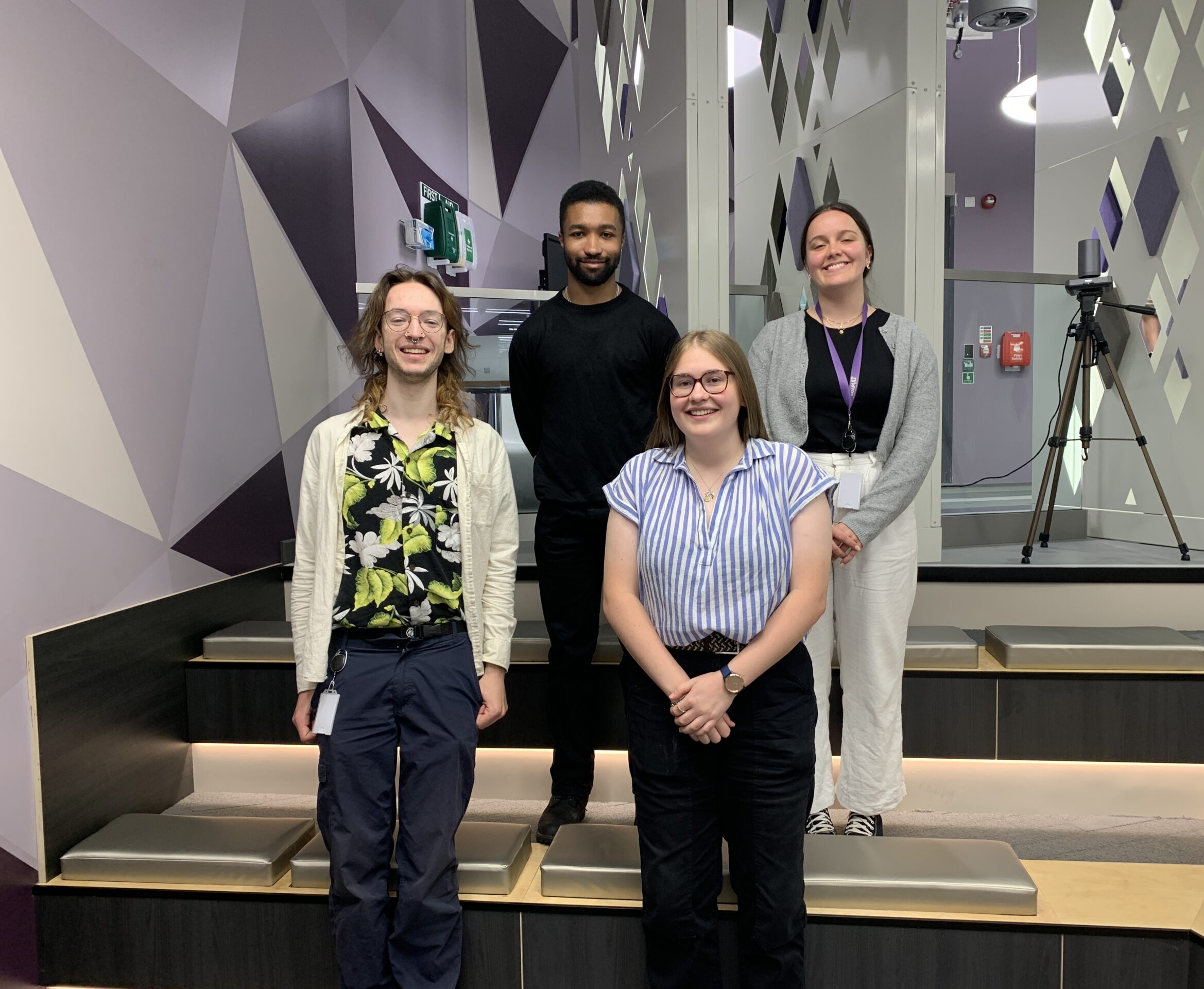 Four people are standing inside a contemporary room with geometric wall designs. They are positioned on slotted steps and are smiling at the camera. There's a tripod with equipment in the background, alongside an eye-catching fire alarm box on the wall. - CSA Catapult