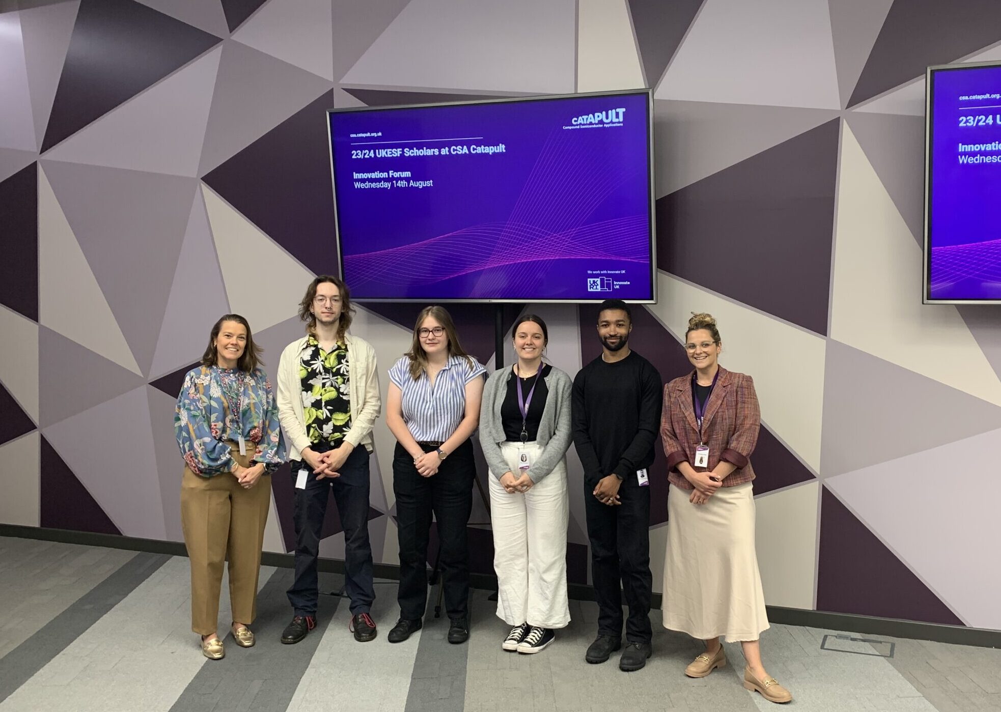 A group of six people stands in a room with geometric patterned walls. Two digital screens behind them display the text: "22/23 UKESF Scholars at CSA Catapult Industry Forum Wednesday 16th August." The group members, smiling and dressed in business casual attire, exude a sense of e-enthusiasm. - CSA Catapult