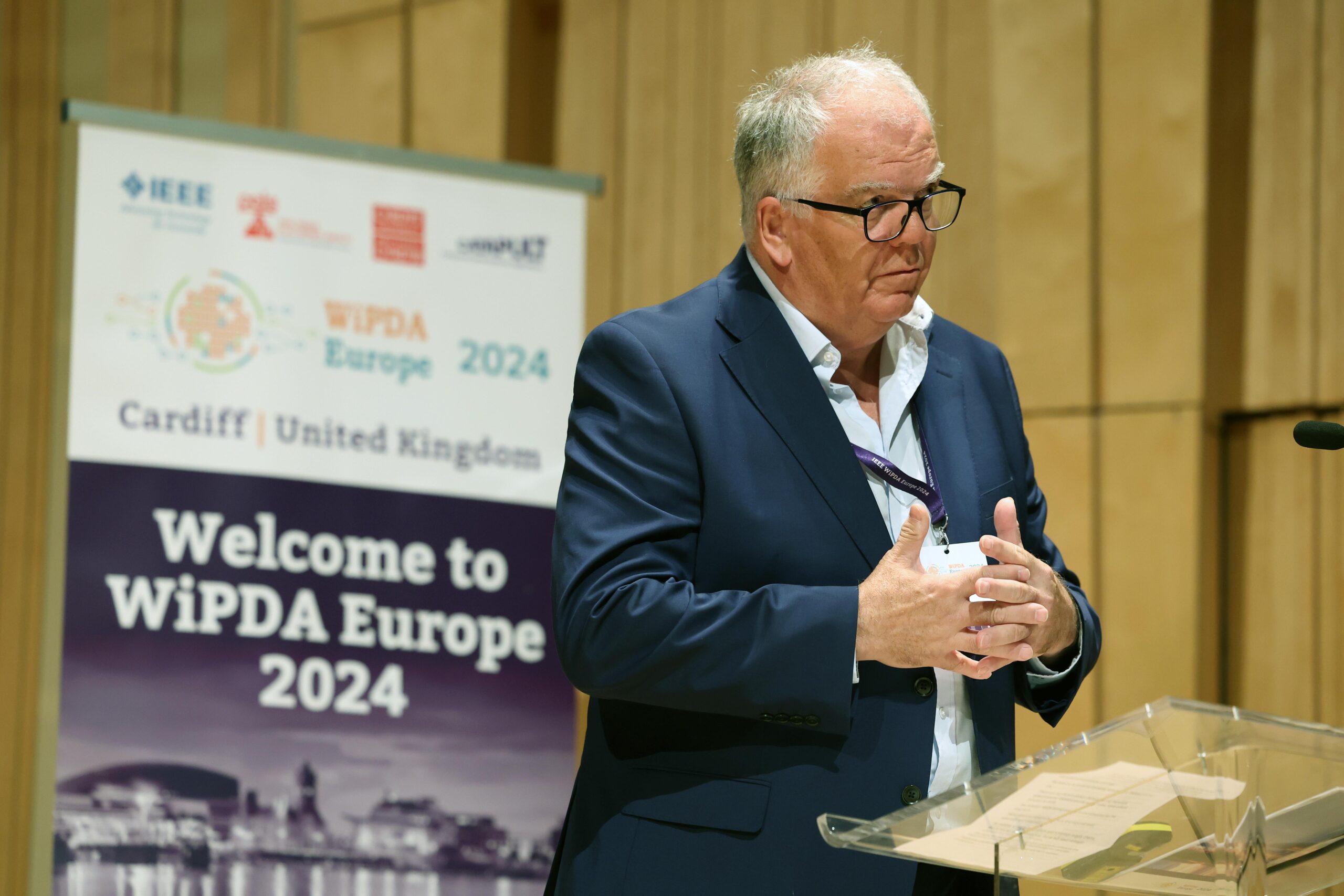 A man in a suit and glasses stands at a podium, speaking at a conference. Behind him is a banner that reads "Welcome to WiPDA Europe 2024" and lists sponsors including IEEE and Cardiff, United Kingdom. The background features wooden panels. - CSA Catapult