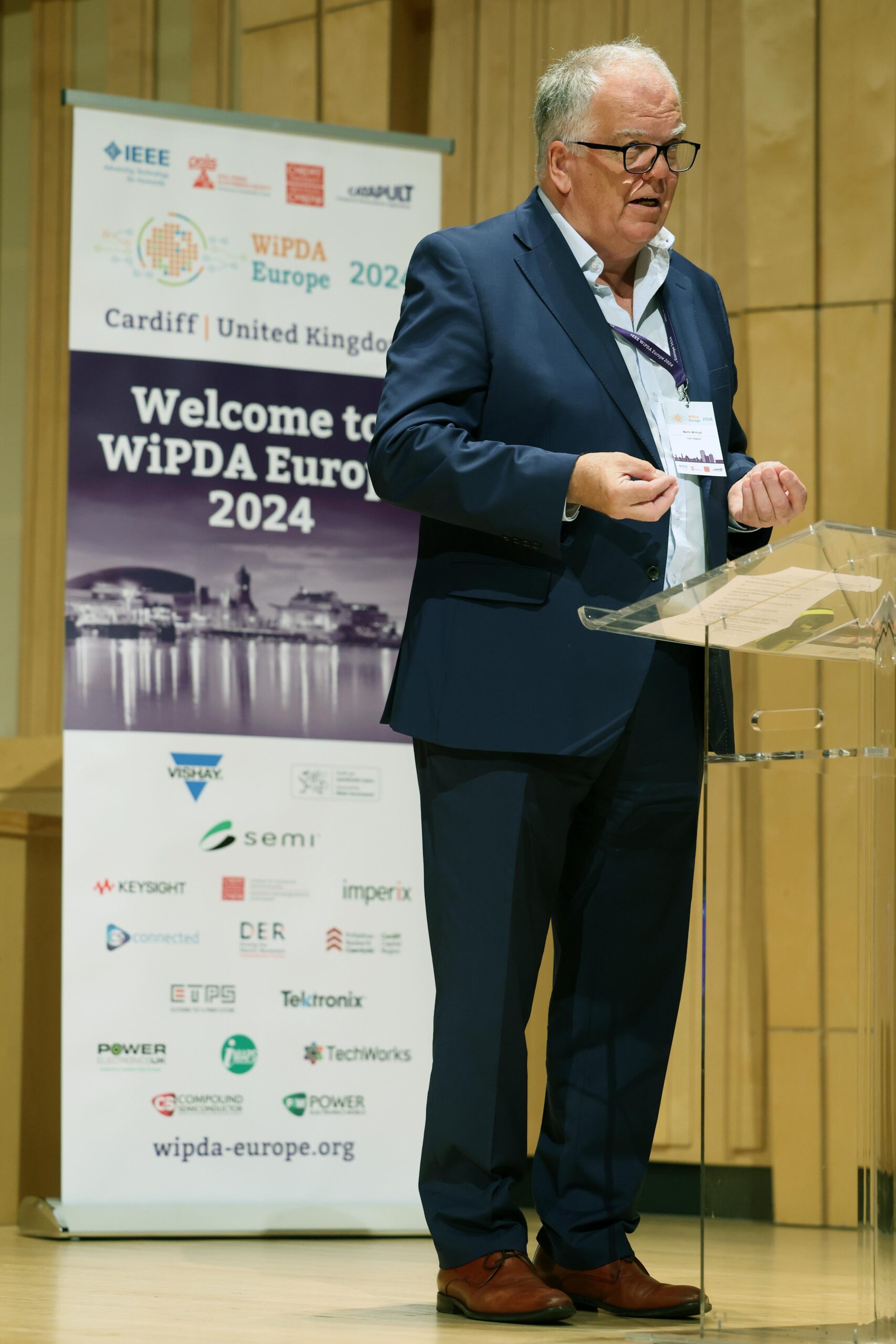 A man in a navy suit stands behind a clear podium, speaking into a microphone. Behind him, there is a banner with the text 