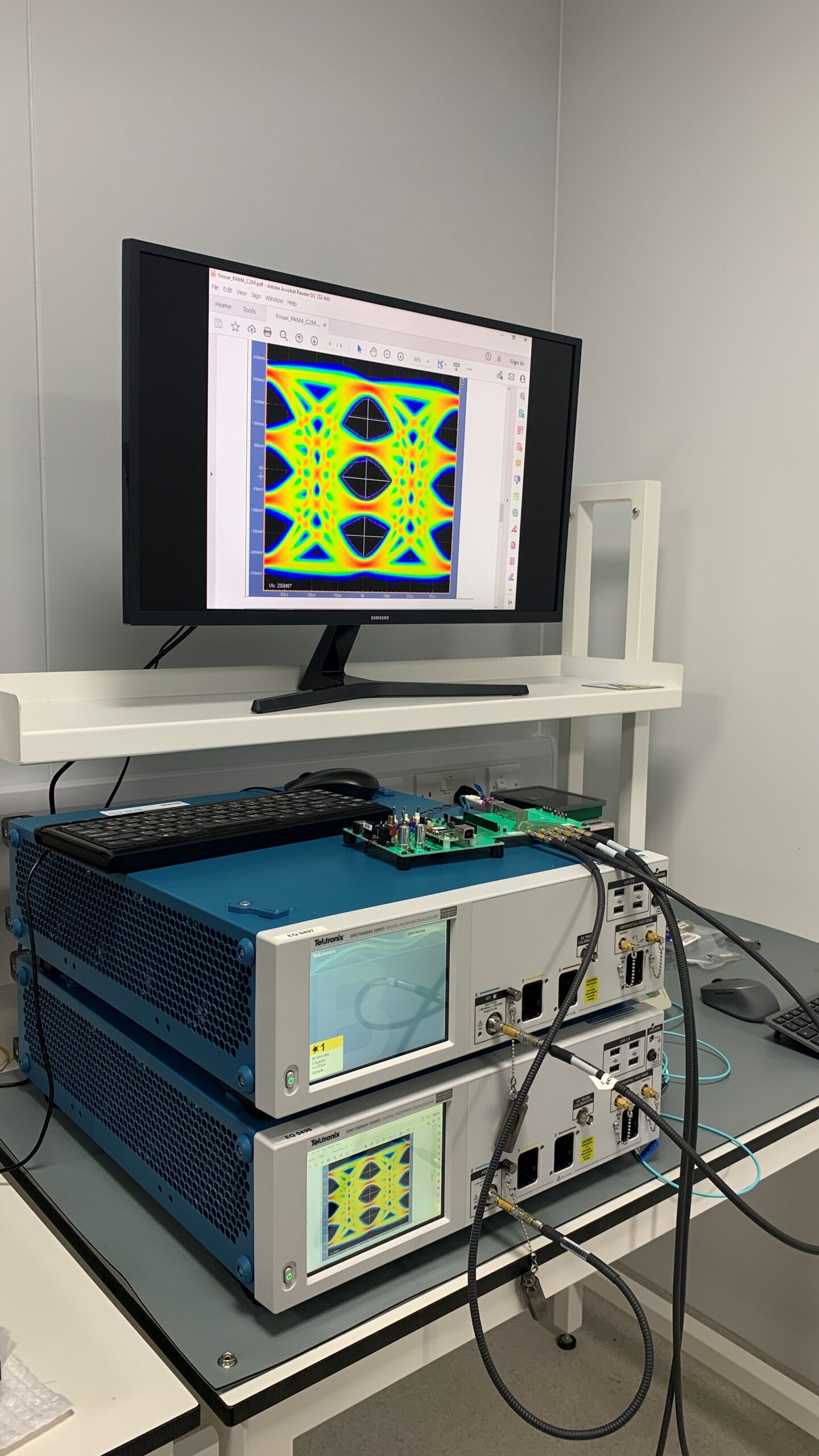 A lab setup with electronic equipment including two blue signal generators connected by cables, a computer monitor displaying a vibrant, symmetrical, multicolored pattern, and a desk with various electronics. The scene suggests testing or calibration of electronic devices using state-of-the-art technology. - CSA Catapult