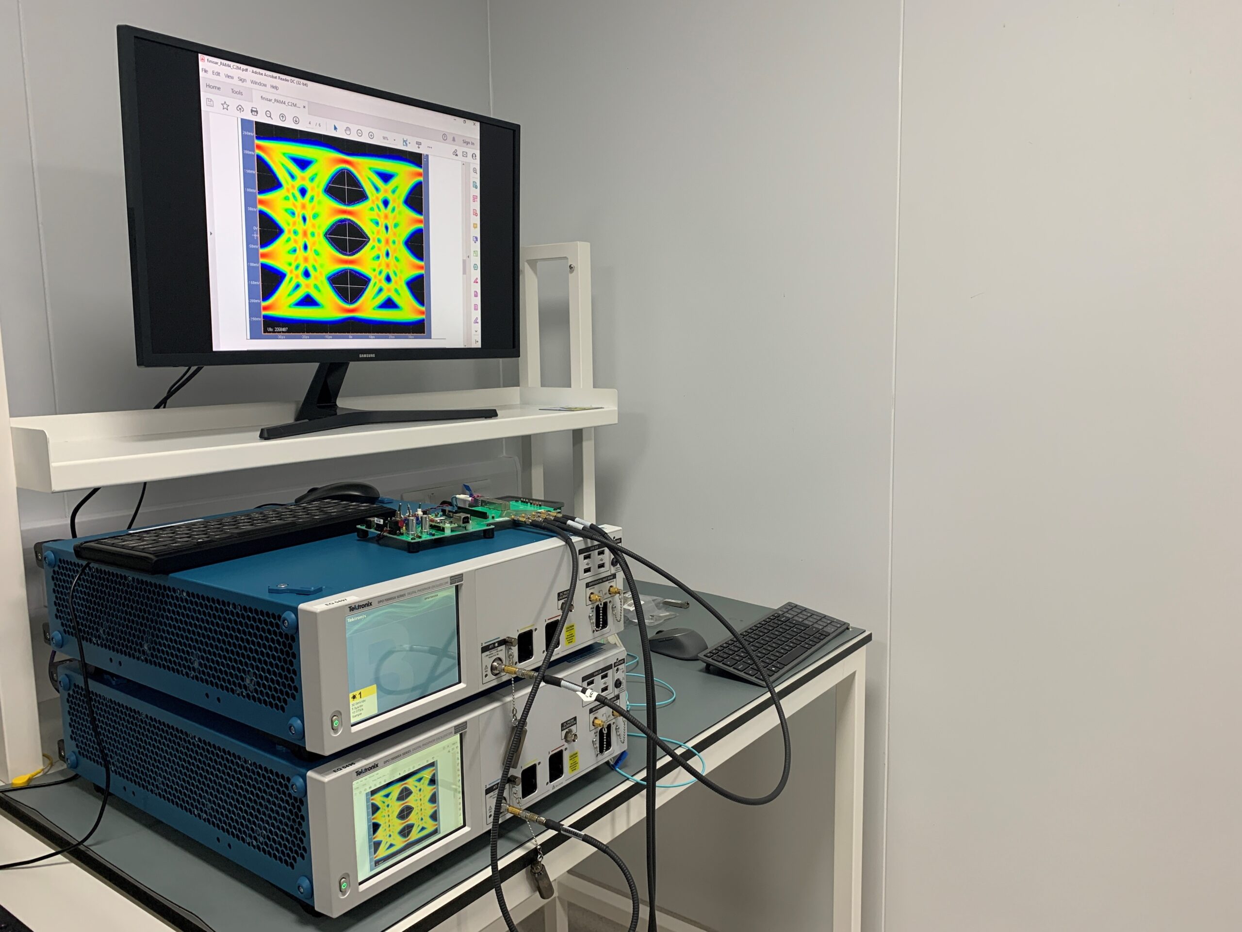 A computer monitor displays a colorful waveform pattern. Below the monitor, two electronic devices with cables connected to a circuit board emit soft hums. A keyboard and a mouse sit on the desk next to the equipment in the clean, white room. - CSA Catapult