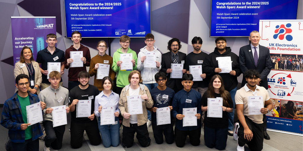 A group of young award winners stands together holding their certificates in front of a backdrop displaying congratulatory messages for the 2024/2025 Welsh Space e-Award winners. They are joined by event organizers. A banner on the right reads "UK Electronics Skills Foundation. - CSA Catapult