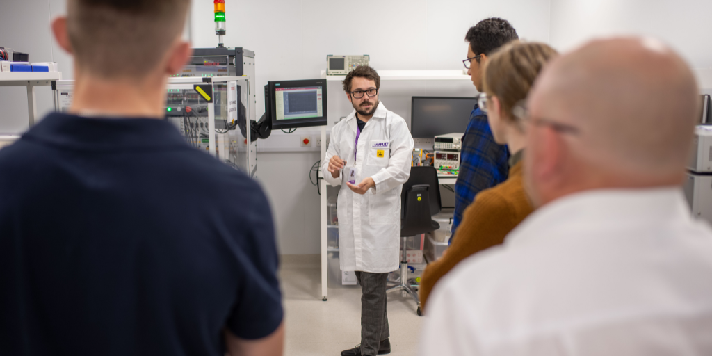 A scientist in a white lab coat explains a concept to a group of four people in a clean, well-lit laboratory. Equipped with various machines and monitors, the lab creates an atmosphere where the individuals listen attentively. - CSA Catapult