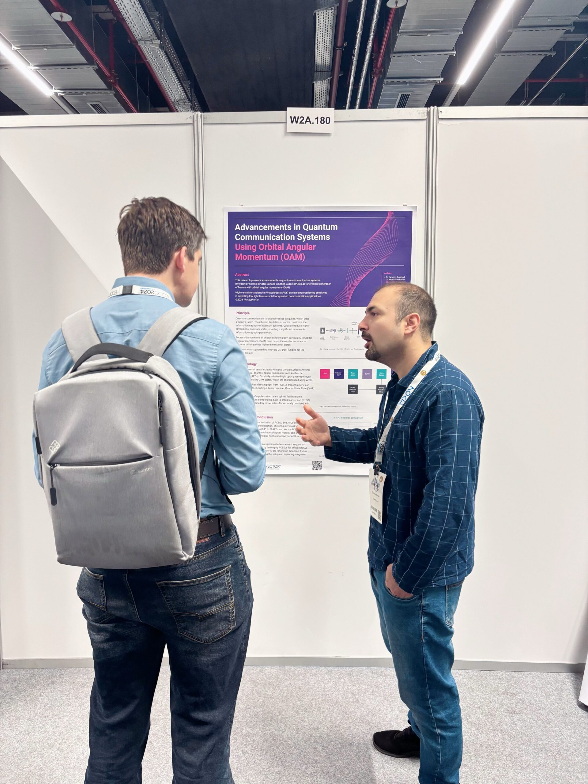 Two men stand in front of a scientific poster at a conference. The man on the left, wearing a backpack, listens attentively. The man on the right, gesturing with his hand, explains the content of the poster titled 