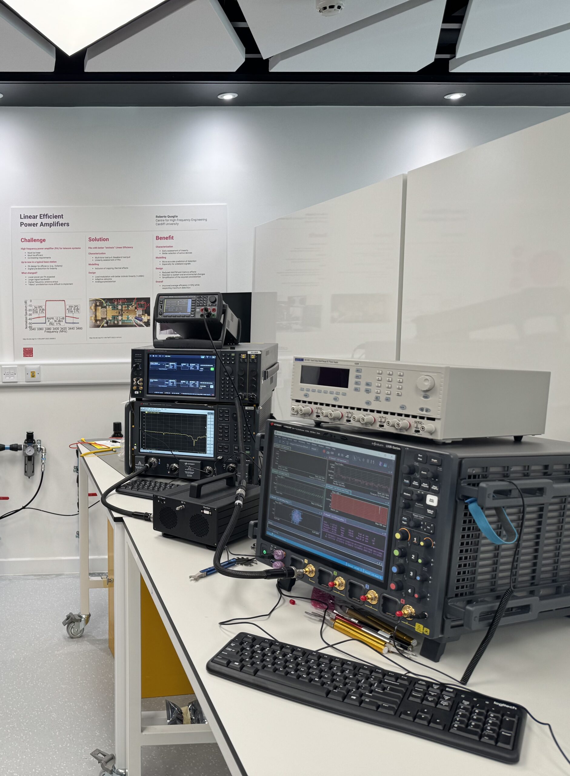 A laboratory with cutting-edge electronic test equipment on a sleek workbench, featuring oscilloscopes and signal generators. In front, a keyboard and mouse invite interaction. A scientific poster brightens the wall of this well-lit, modern e-engineering space. - CSA Catapult