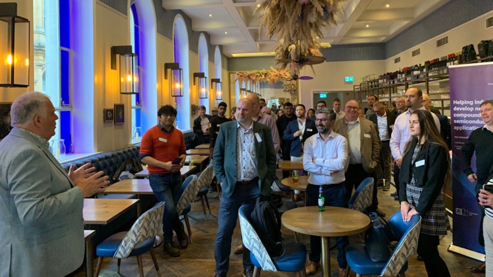 A man is speaking to a group of people gathered in a modern, well-lit room with large windows and decorative hanging elements. Attendees, dressed in business casual attire, are standing or sitting on chairs, attentively listening. Tables are arranged around the room e - CSA Catapult