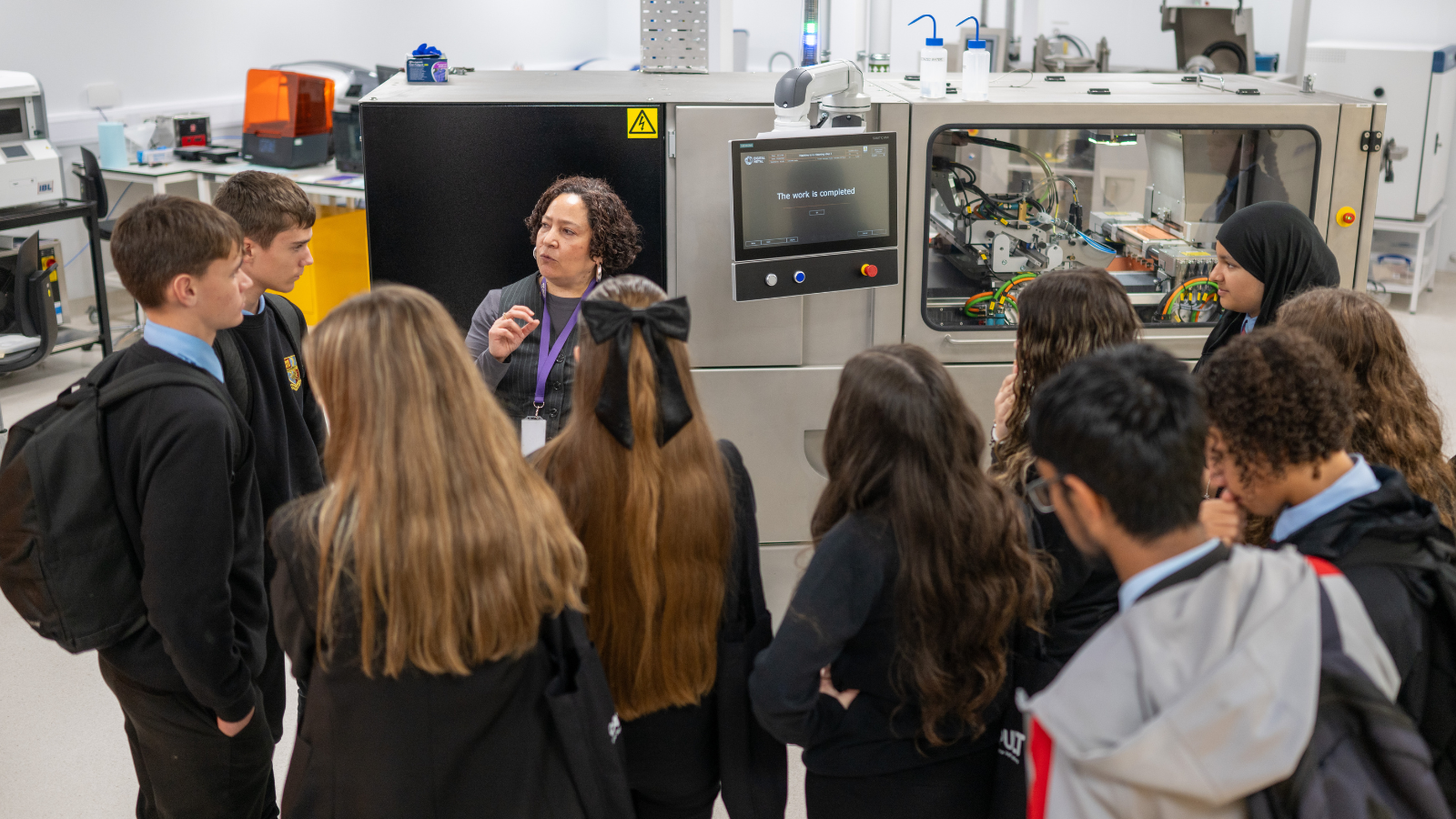 A group of eager students listens intently to a presenter in a laboratory setting, with advanced technical equipment and electronic screens glowing softly in the background. - CSA Catapult