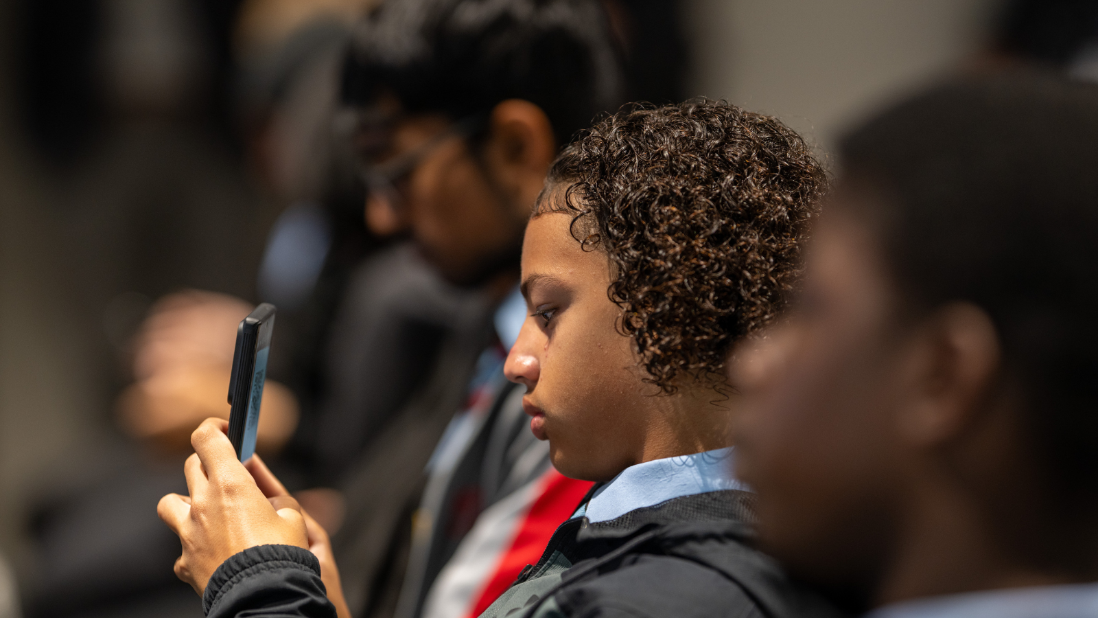 A person with curly hair is engrossed in their phone, seated among others in a lively row of activity. - CSA Catapult