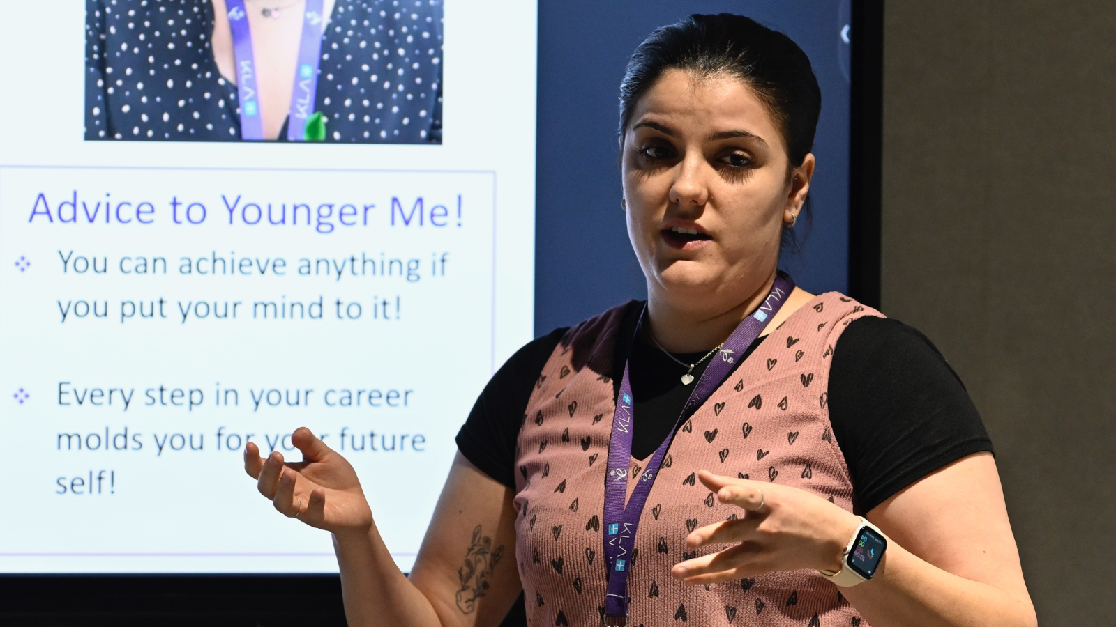 A woman in a patterned vest energetically speaks, e-learning clicker in hand, in front of a presentation slide titled 