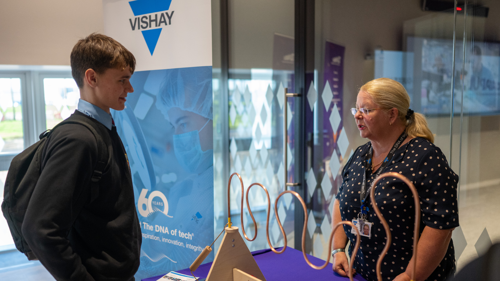 At a bustling event, a woman at the Vishay booth expertly presents electronic components to an eager young man. - CSA Catapult