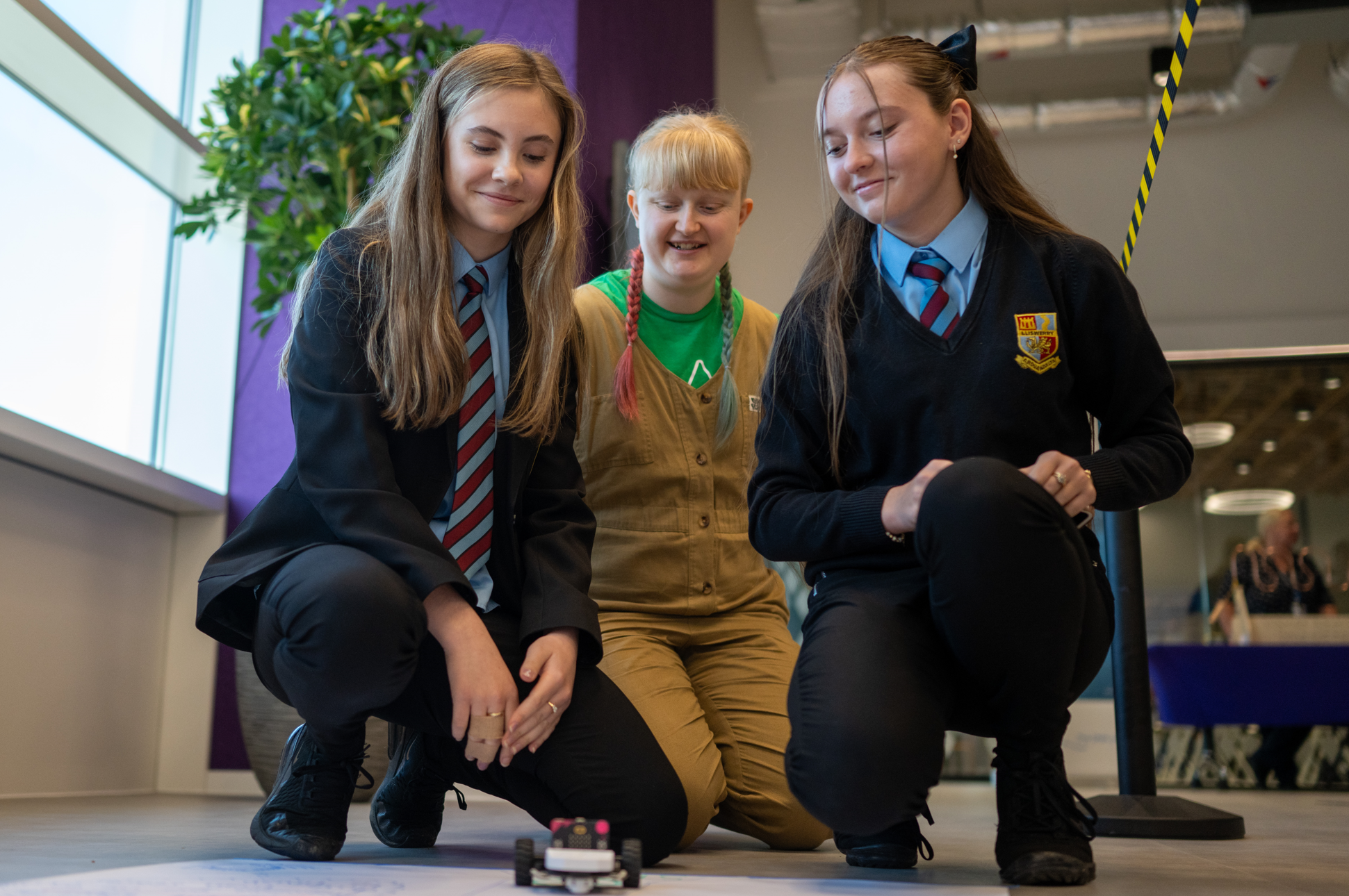 Three enthusiastic young people in uniforms eagerly observe a petite robot navigating the floor indoors. - CSA Catapult