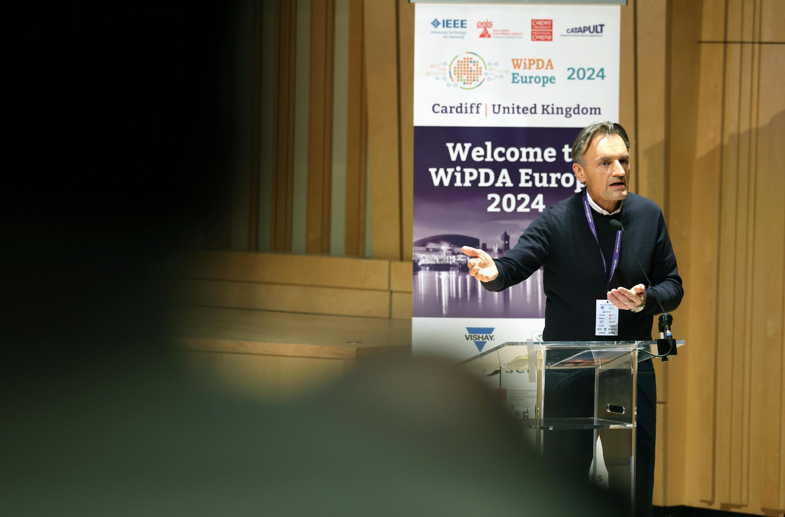 Speaker addressing an audience at WiPDA Europe 2024 in Cardiff, at a conference podium with event banners in the background. - CSA Catapult