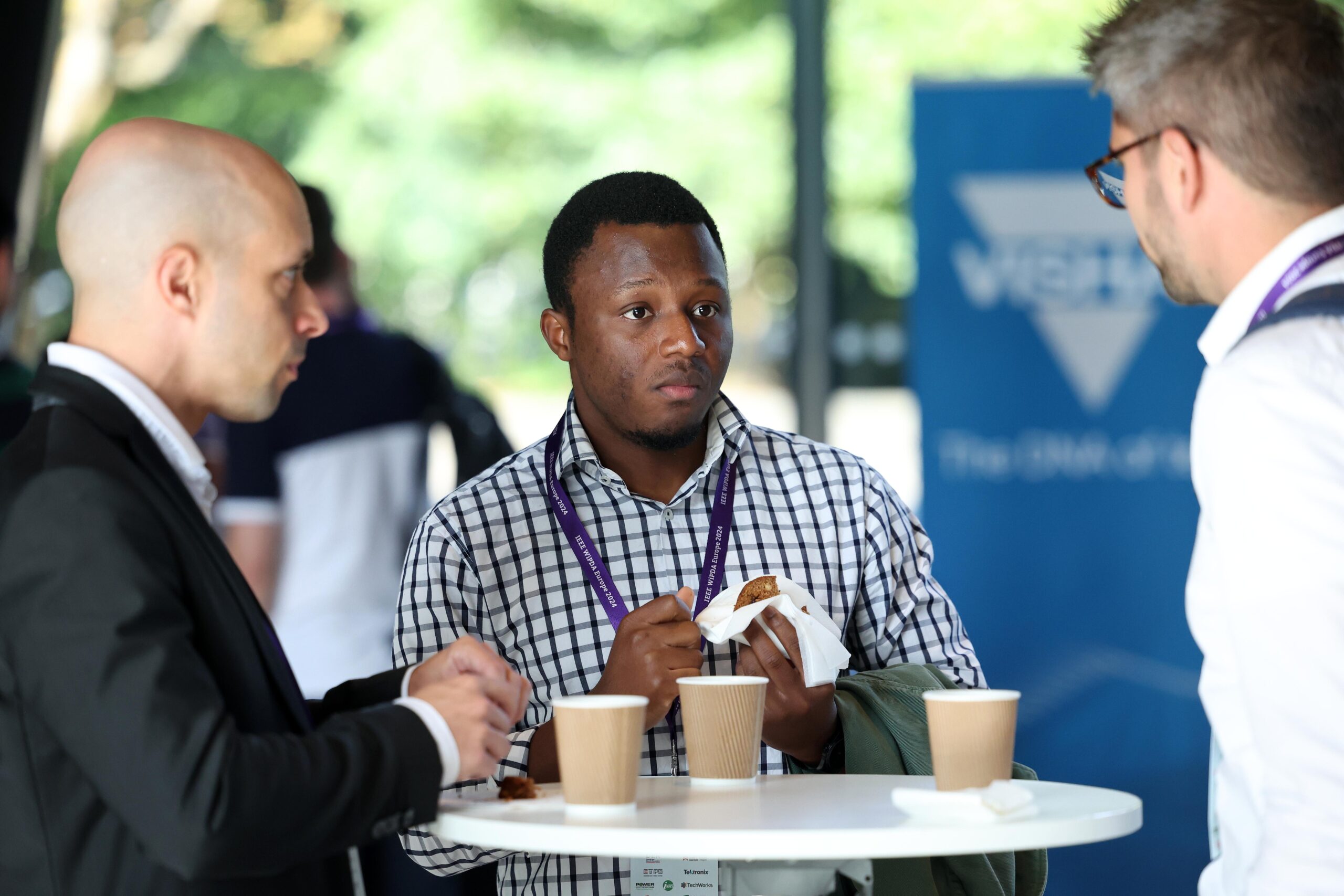 Three men engage in conversation around a standing table with coffee cups and snacks. - CSA Catapult