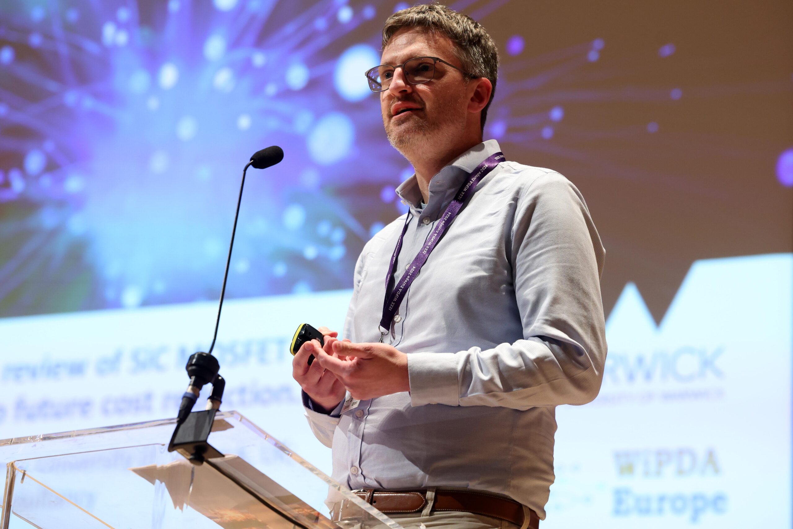 A person speaking at a podium during a conference, with a screen displaying text and logos in the background. - CSA Catapult