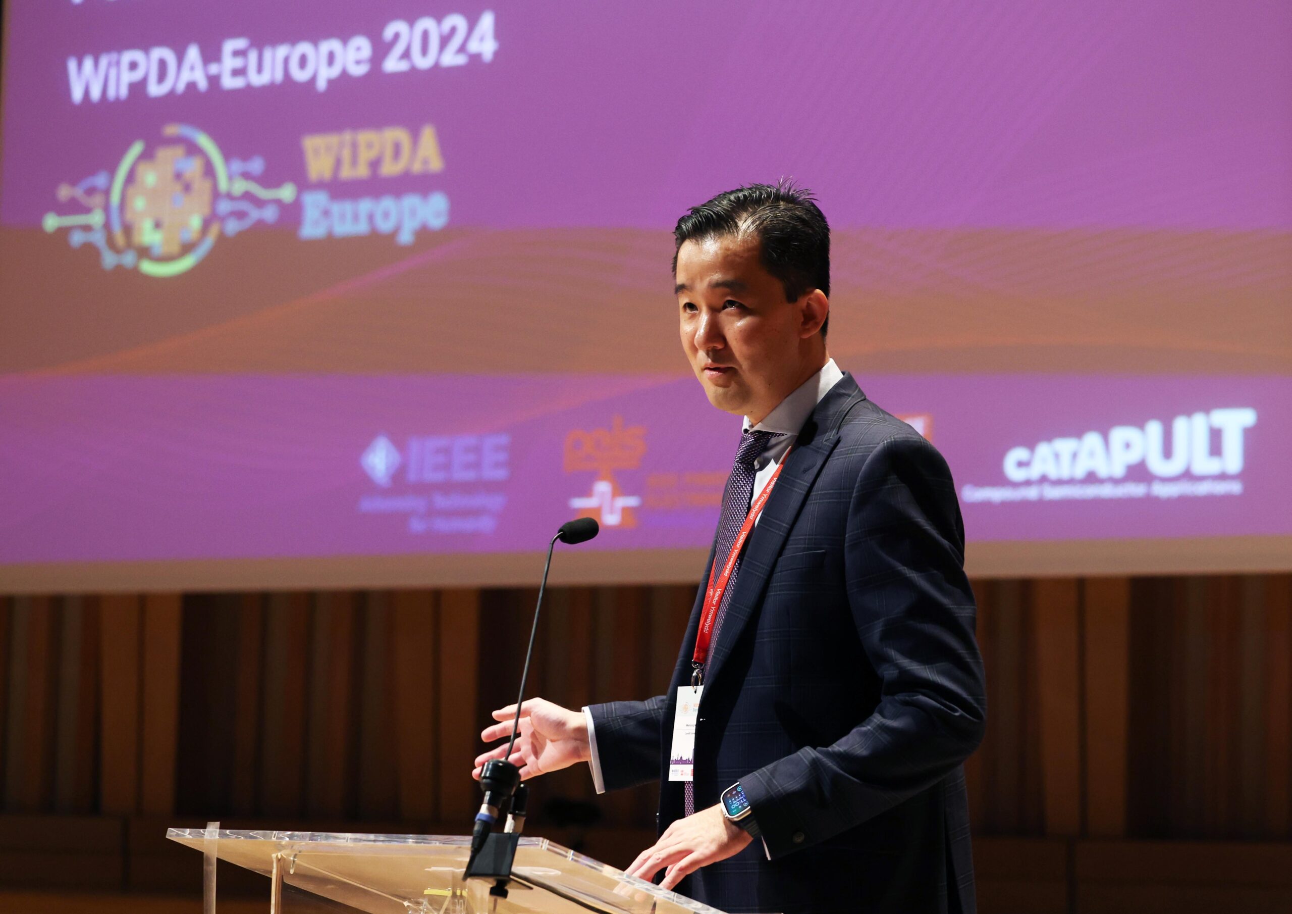 A man speaking at a podium during the WiPDA-Europe 2024 conference, with a purple presentation screen in the background. - CSA Catapult