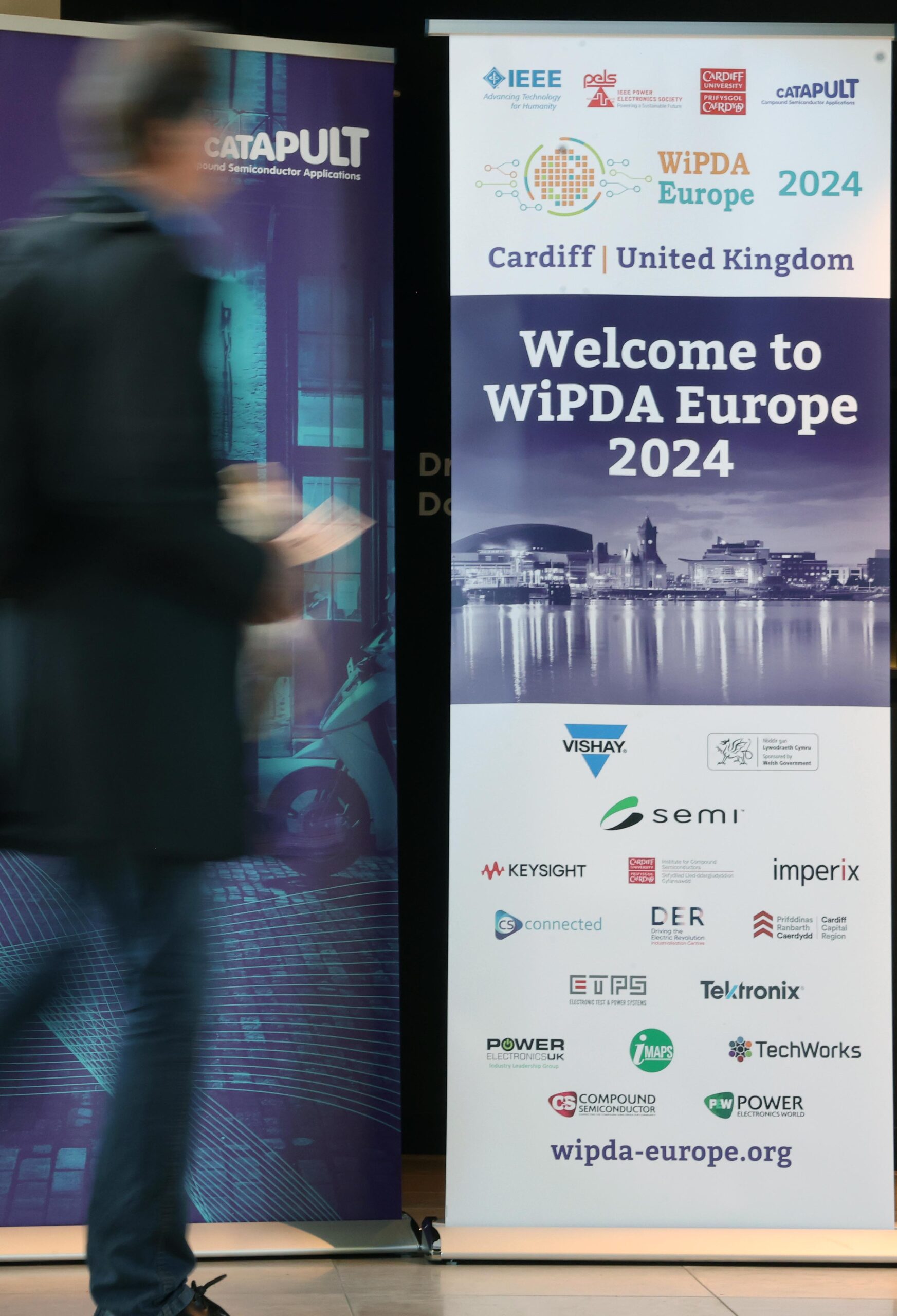 A person walks past banners for WiPDA Europe 2024 in Cardiff, United Kingdom, with logos of sponsors and collaborators. - CSA Catapult