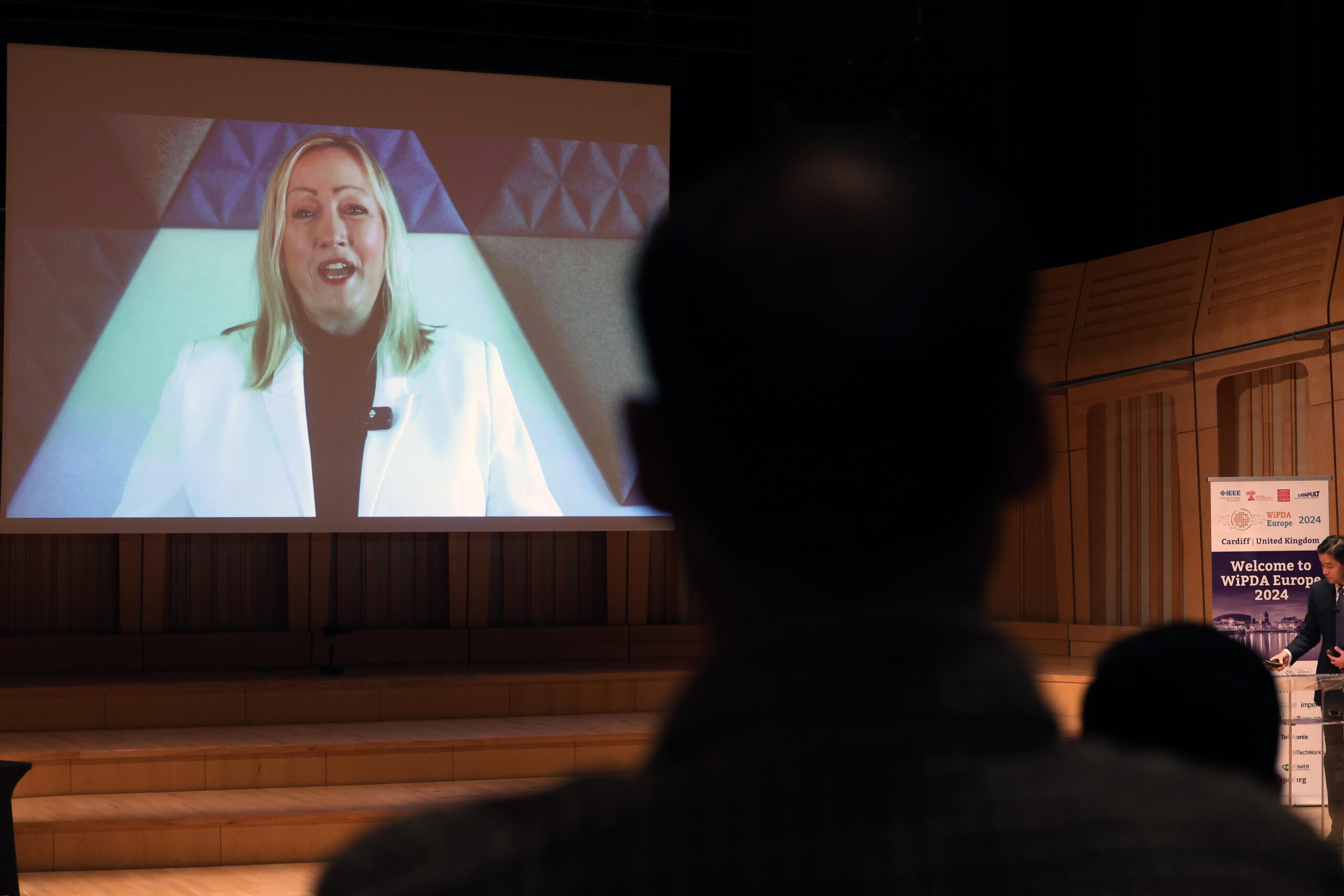 A person watches a woman giving a presentation on a large screen in a dimly lit auditorium. - CSA Catapult