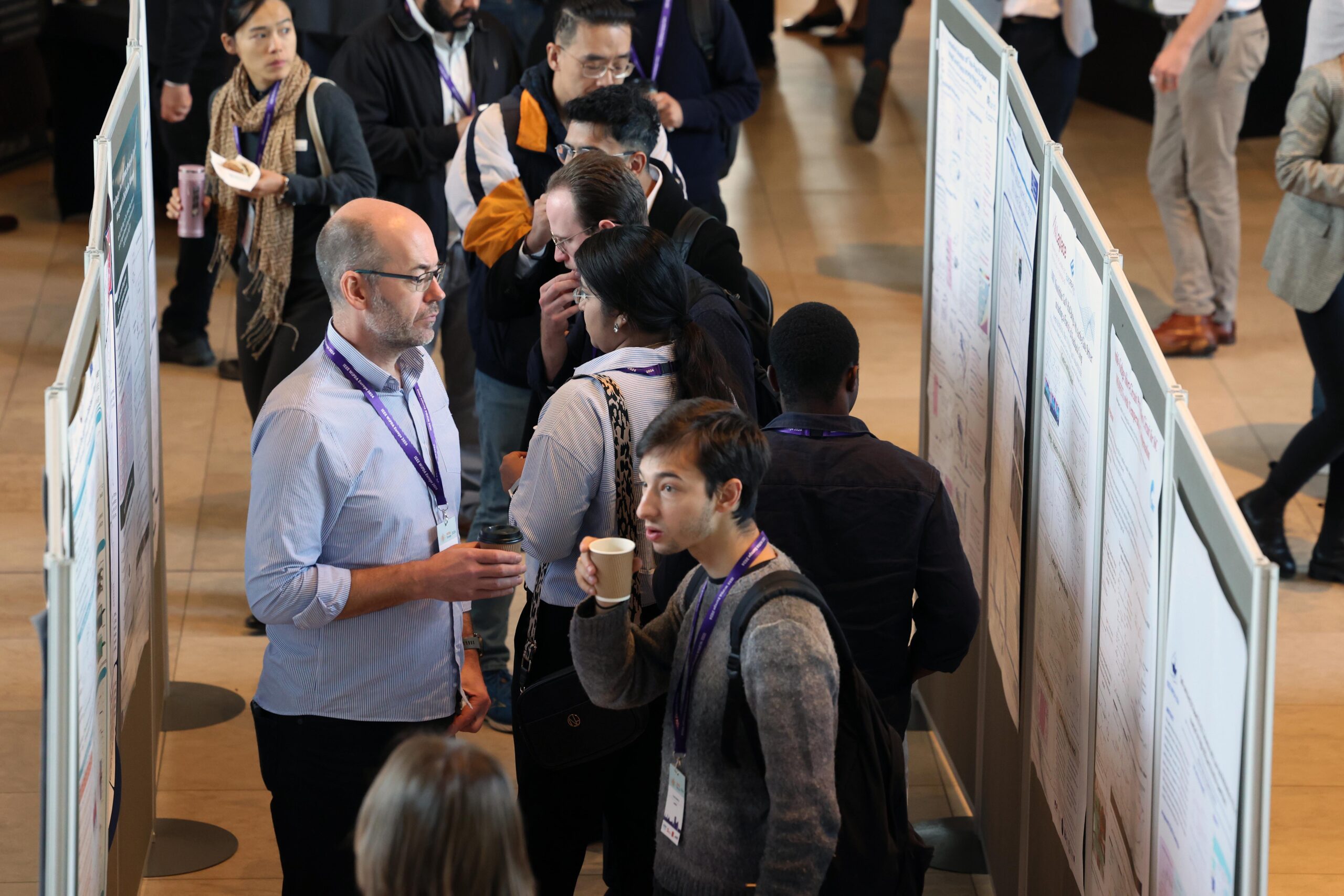 Attendees discussing posters at a conference, some holding coffee cups, in a crowded exhibition space. - CSA Catapult