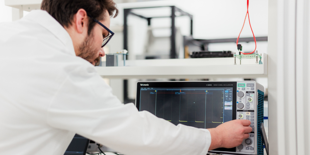 A scientist in a lab coat adjusts settings on a digital oscilloscope displaying waveforms. - CSA Catapult