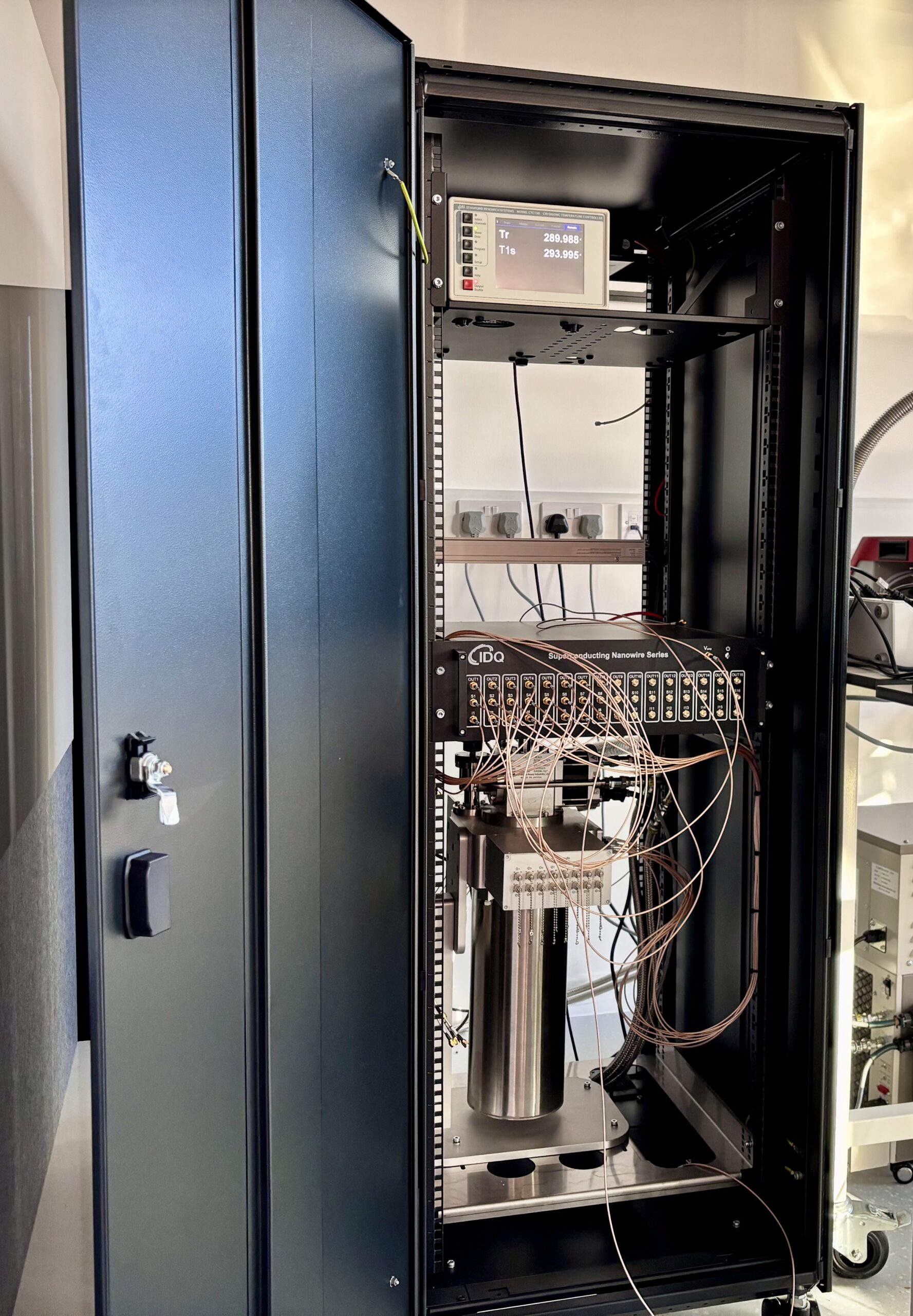 Cabinet housing a quantum computer setup, featuring a central cylindrical component and connected wires, with a control panel above. - CSA Catapult