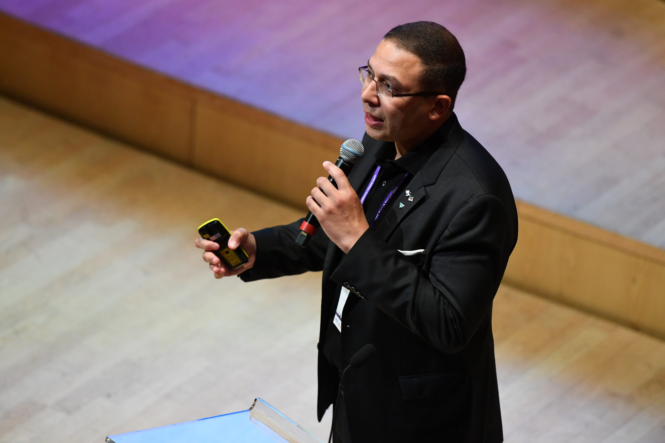 Man in a suit speaking into a microphone on stage, holding a smartphone, with a wooden floor background. - CSA Catapult