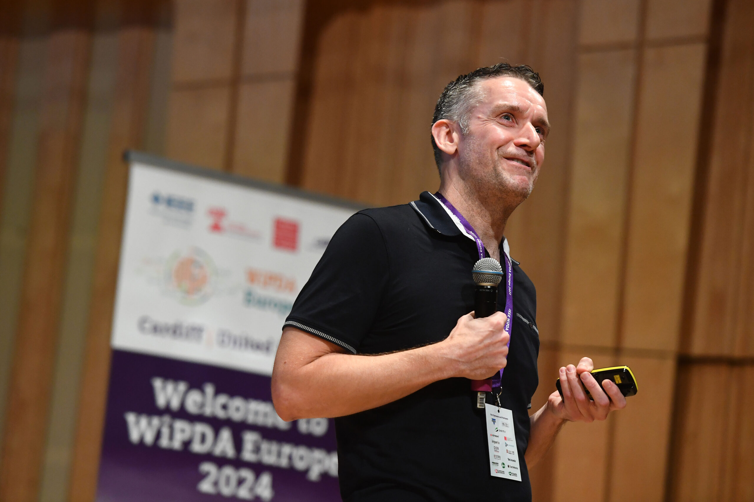 A man holds a microphone and a device, standing in front of a sign that reads 