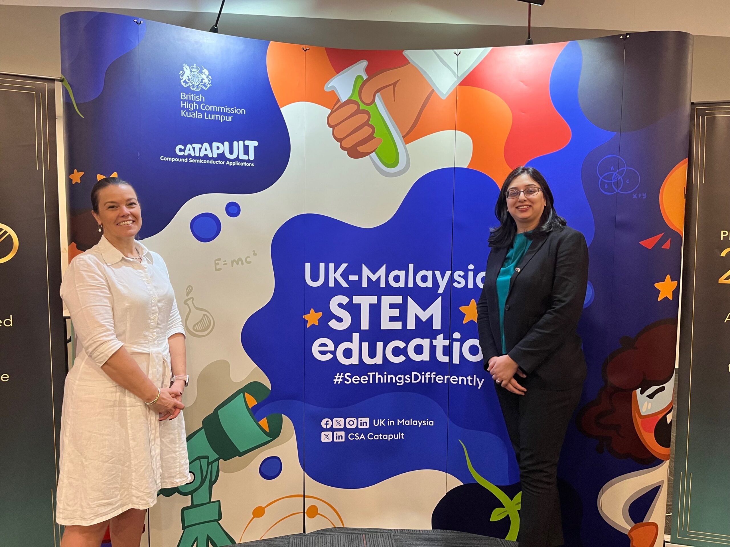 Two women pose before a UK-Malaysia STEM education banner, showcasing e-learning innovations with scientific illustrations. - CSA Catapult