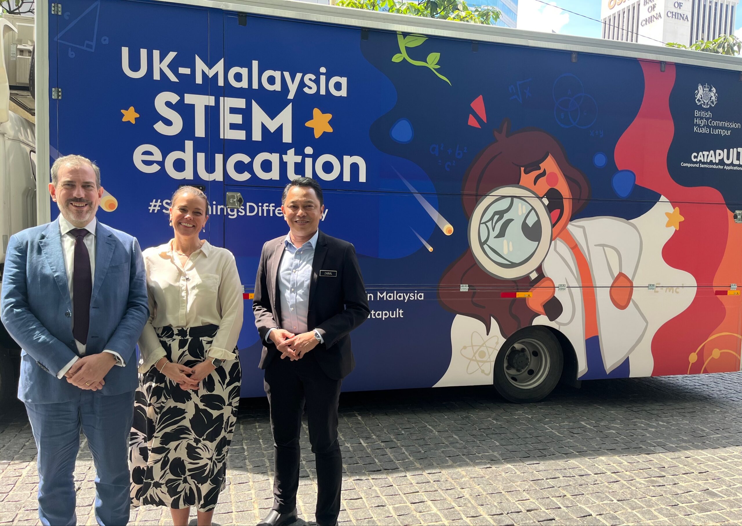 Three people pose by a vibrant UK-Malaysia STEM education van featuring an illustration of a curious scientist. - CSA Catapult