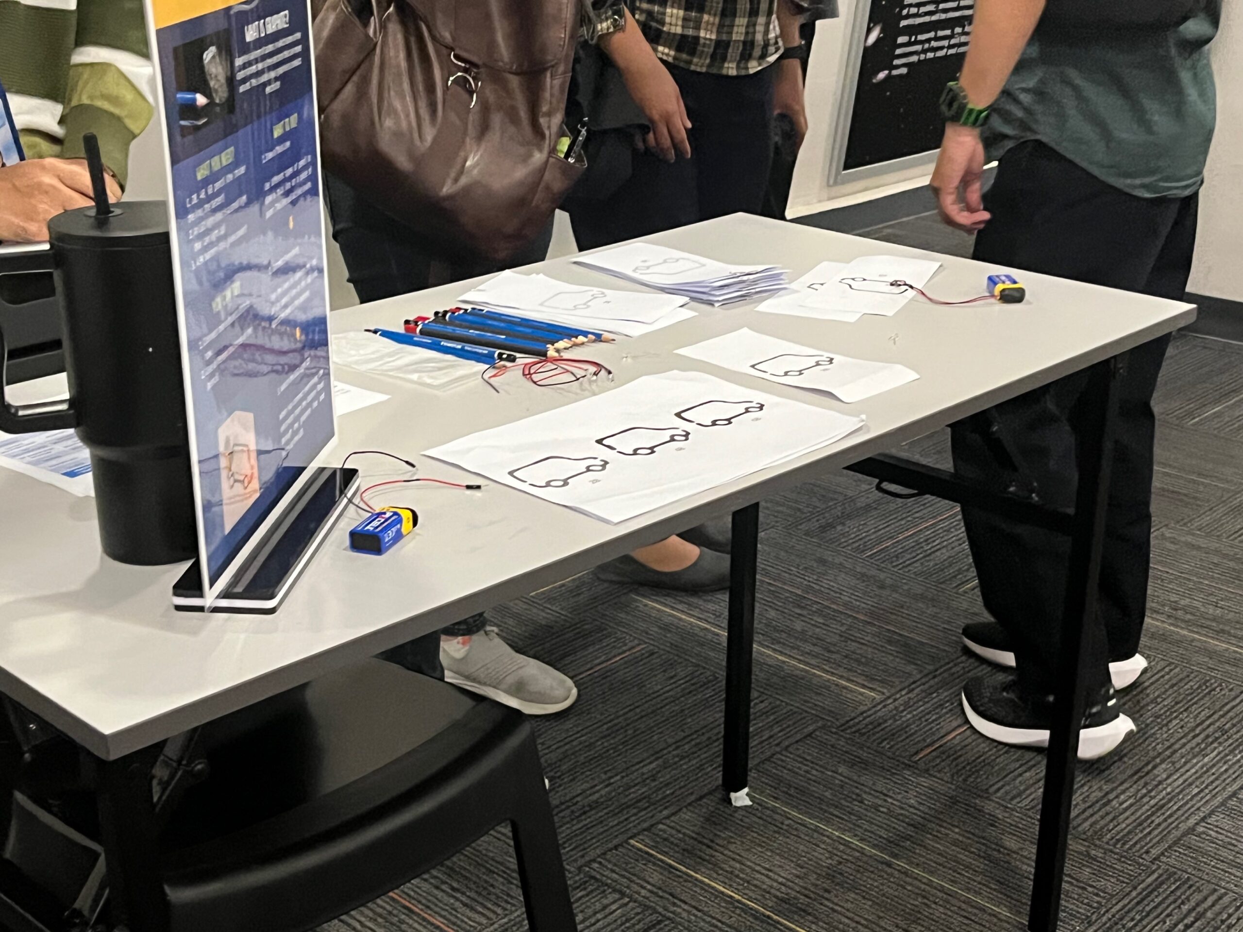 Table with electronic components, e-papers with circuit designs, and people standing nearby. - CSA Catapult