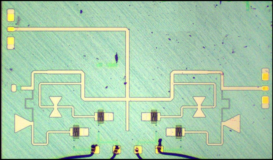 Close-up of a microchip with a circuit design on a blue surface, featuring pathways and components arranged symmetrically. - CSA Catapult