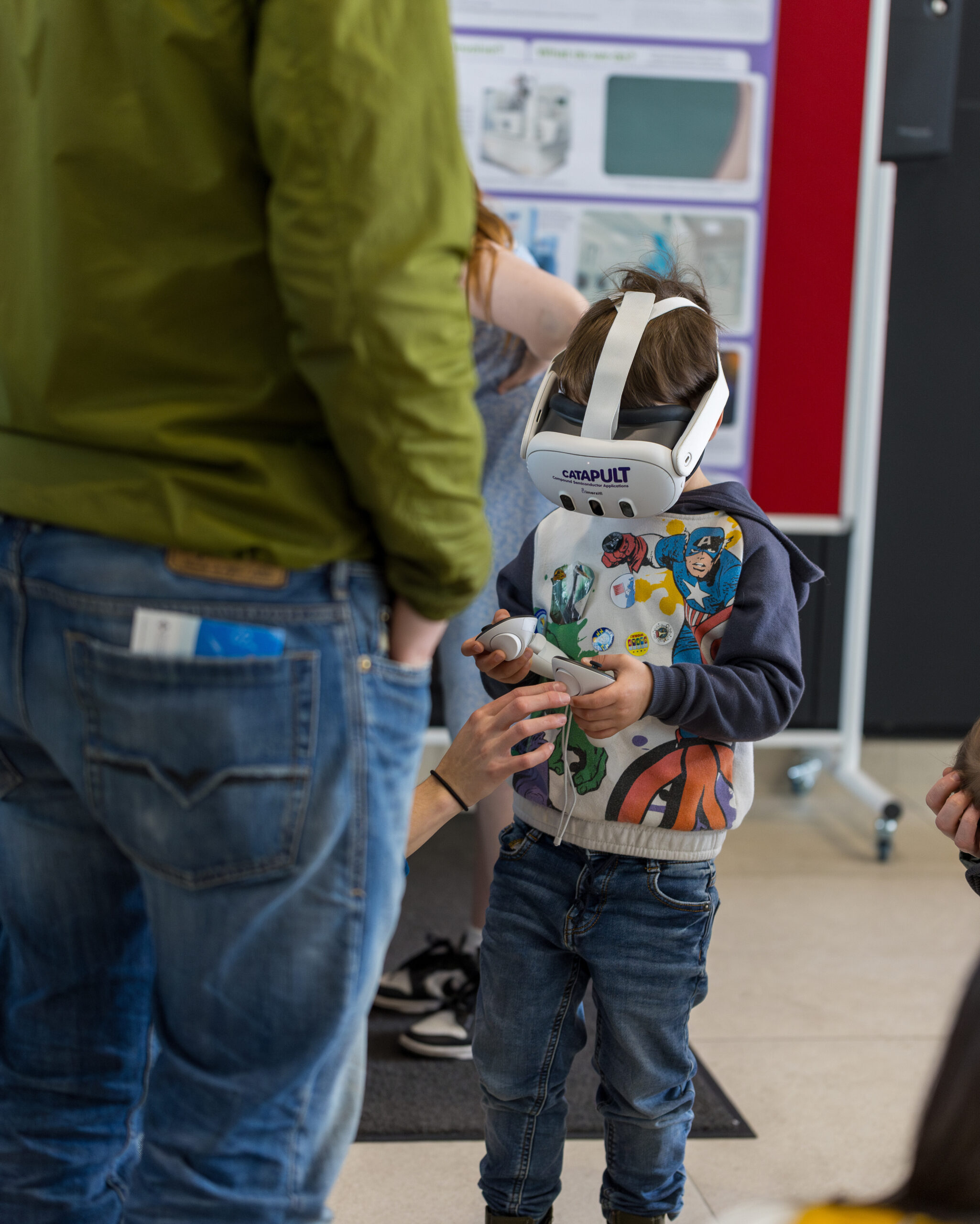 At the Science Festival, a child dons a VR headset, guided by an adult, while others watch near vibrant posters. - CSA Catapult