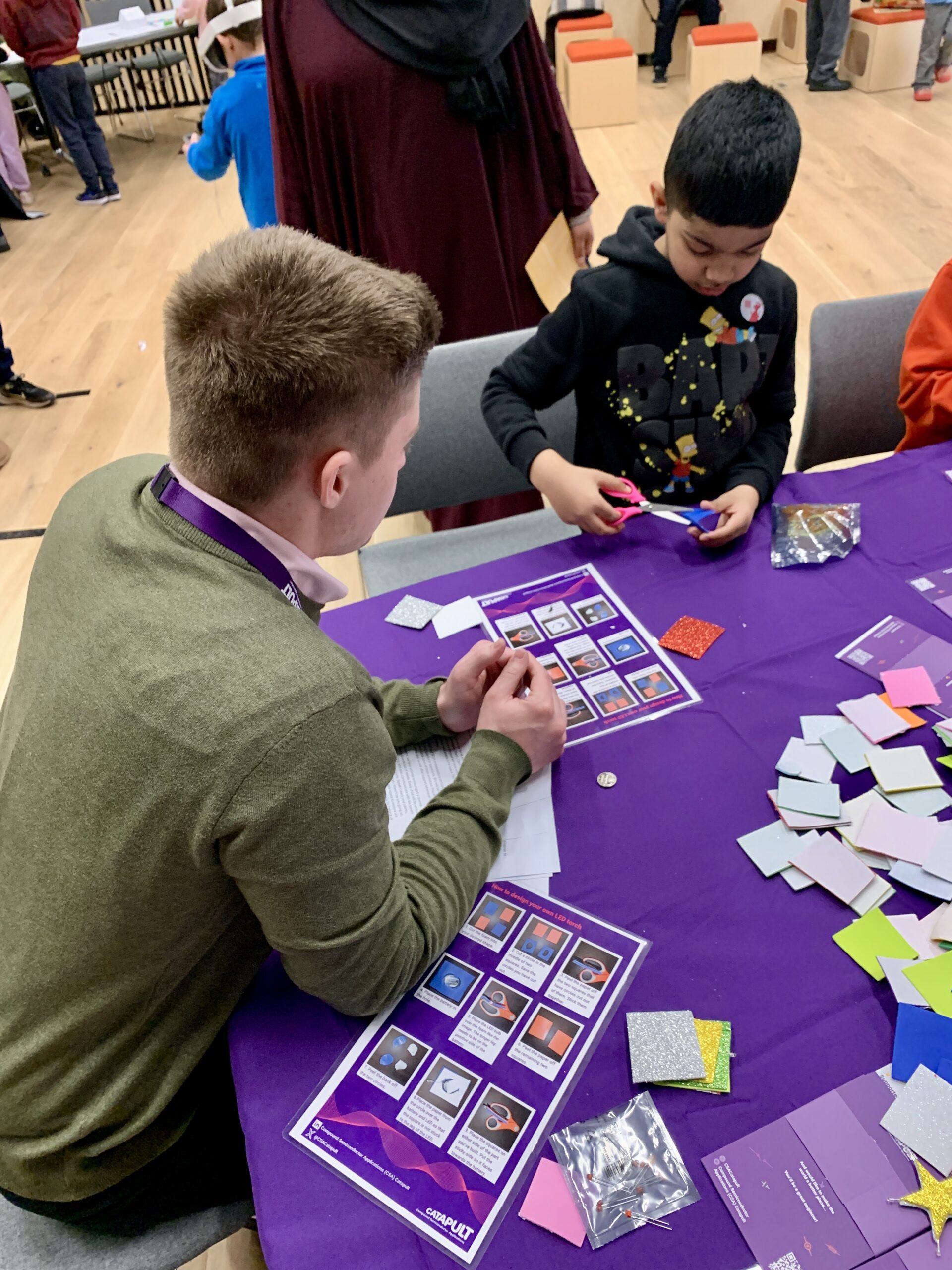 An adult and child engage in a crafting activity at a CSA Skills Academy table filled with colored paper and craft materials. - CSA Catapult