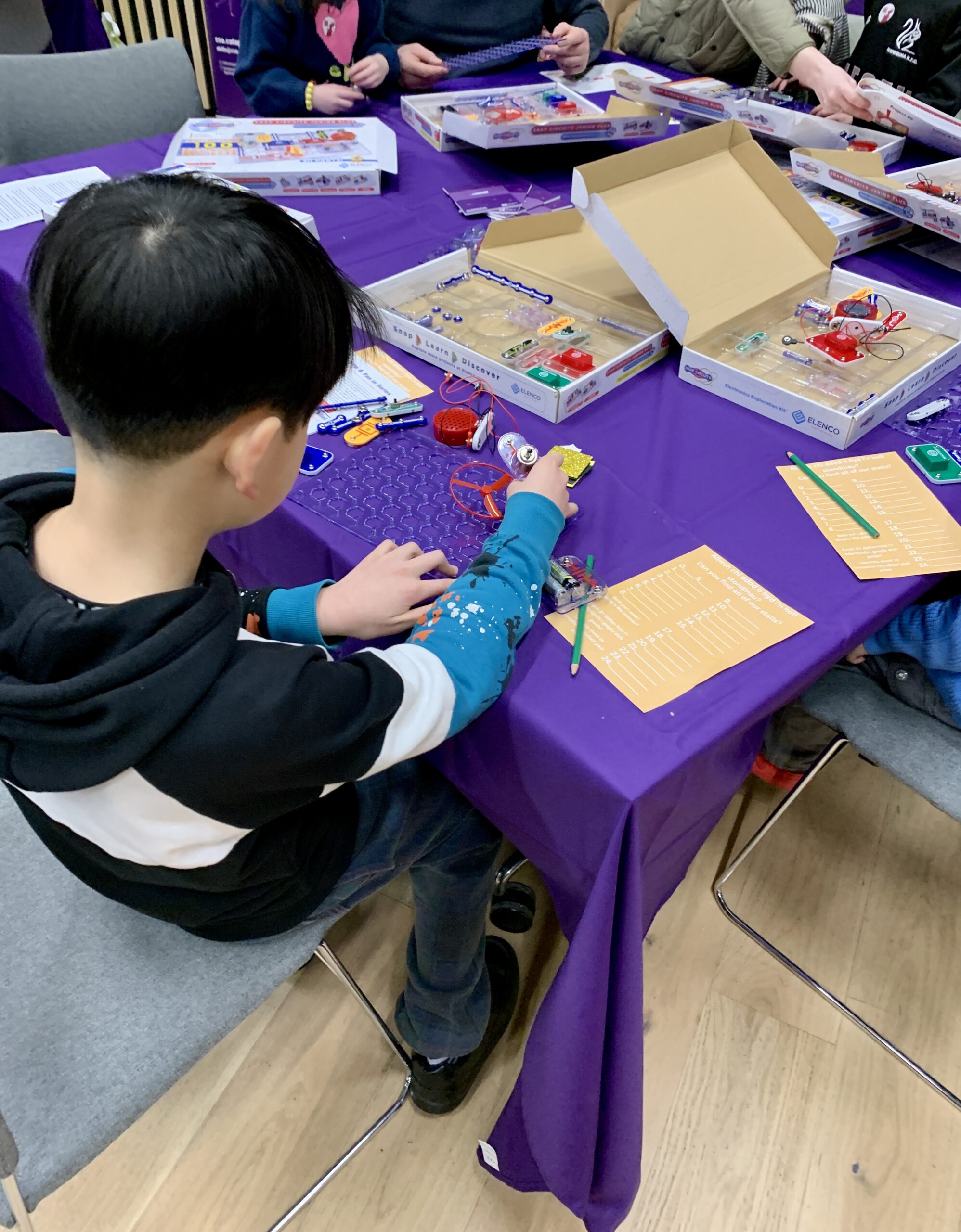 A child at the CSA Catapult Skills Academy table at Be A Scientist explores electronics on a purple tablecloth, surrounded by components and guides. - CSA Catapult