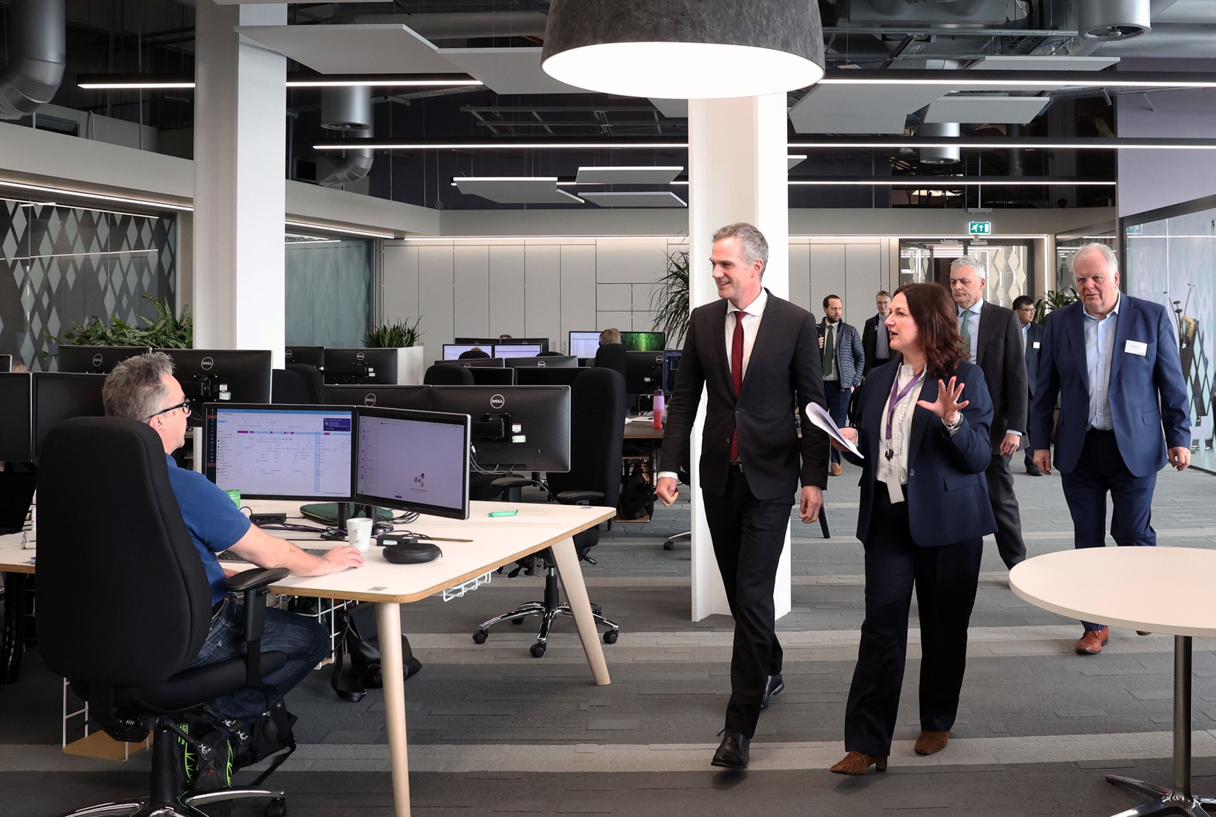 A group in business attire tours a modern AME office with multiple workstations and a large overhead lamp. - CSA Catapult