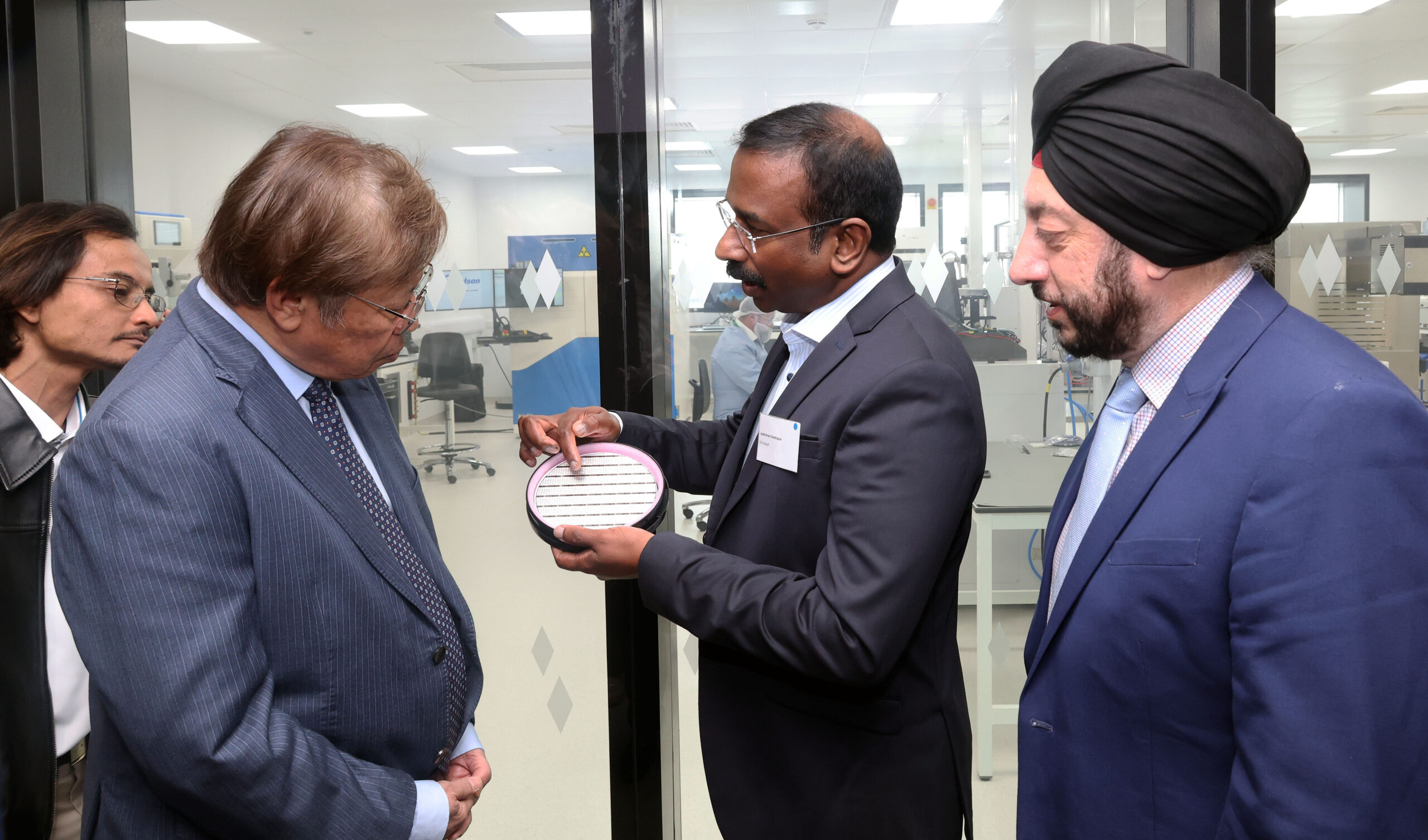 A man shows a silicon wafer to two others in suits during a Sarawak lab tour. - CSA Catapult